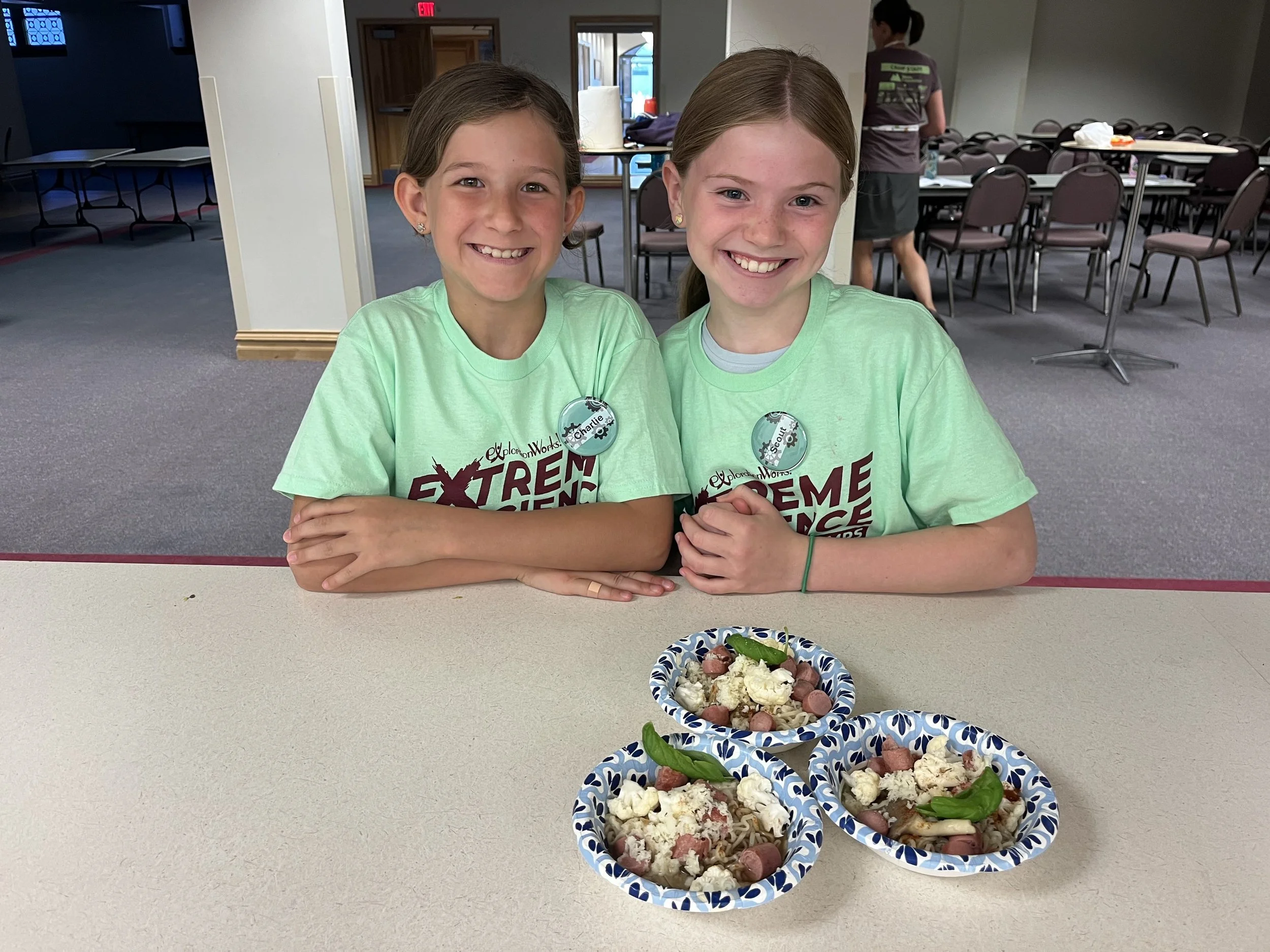 Two young girls wearing matching green 'Extreme' T-shirts and badges sit at a table with three bowls of salad topped with vegetables and cheese, smiling at the camera in a community room.