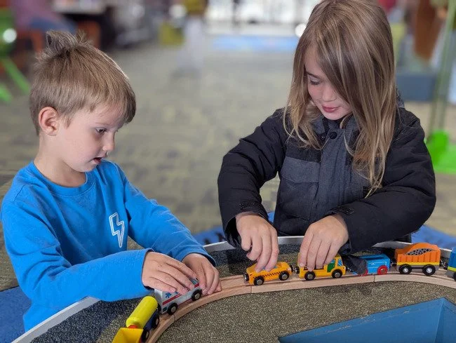Two children playing with toy train set on a circular track outdoors.
