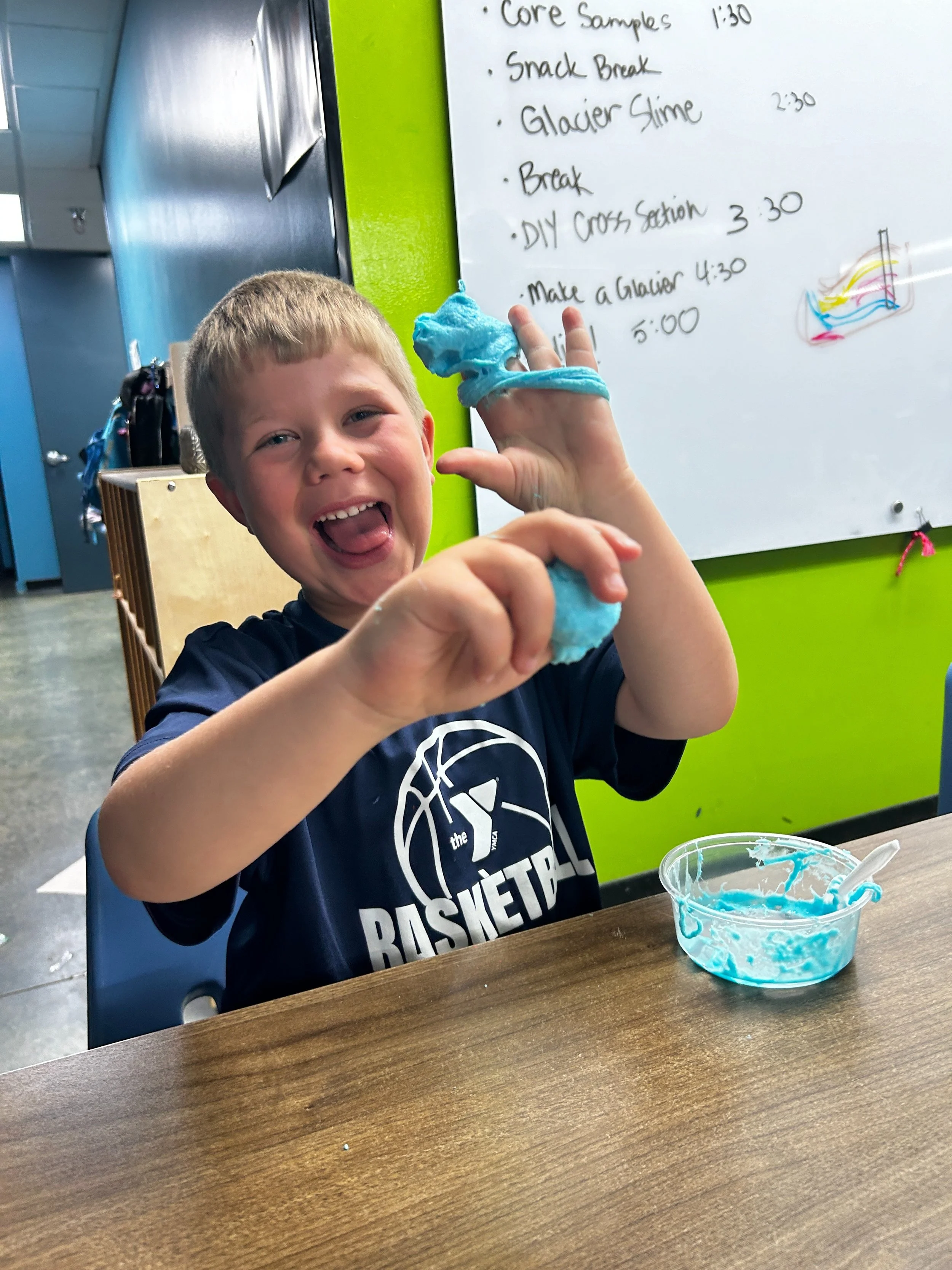 A young boy with a big smile, wearing a sports jersey, is playing with blue slime at a table. He is holding a handful of slime in his right hand and has some slime on his left hand. A small clear plastic bowl with blue slime and a white spatula is on the table in front of him. In the background, there is a whiteboard with a schedule written on it, and the wall behind him is painted green.