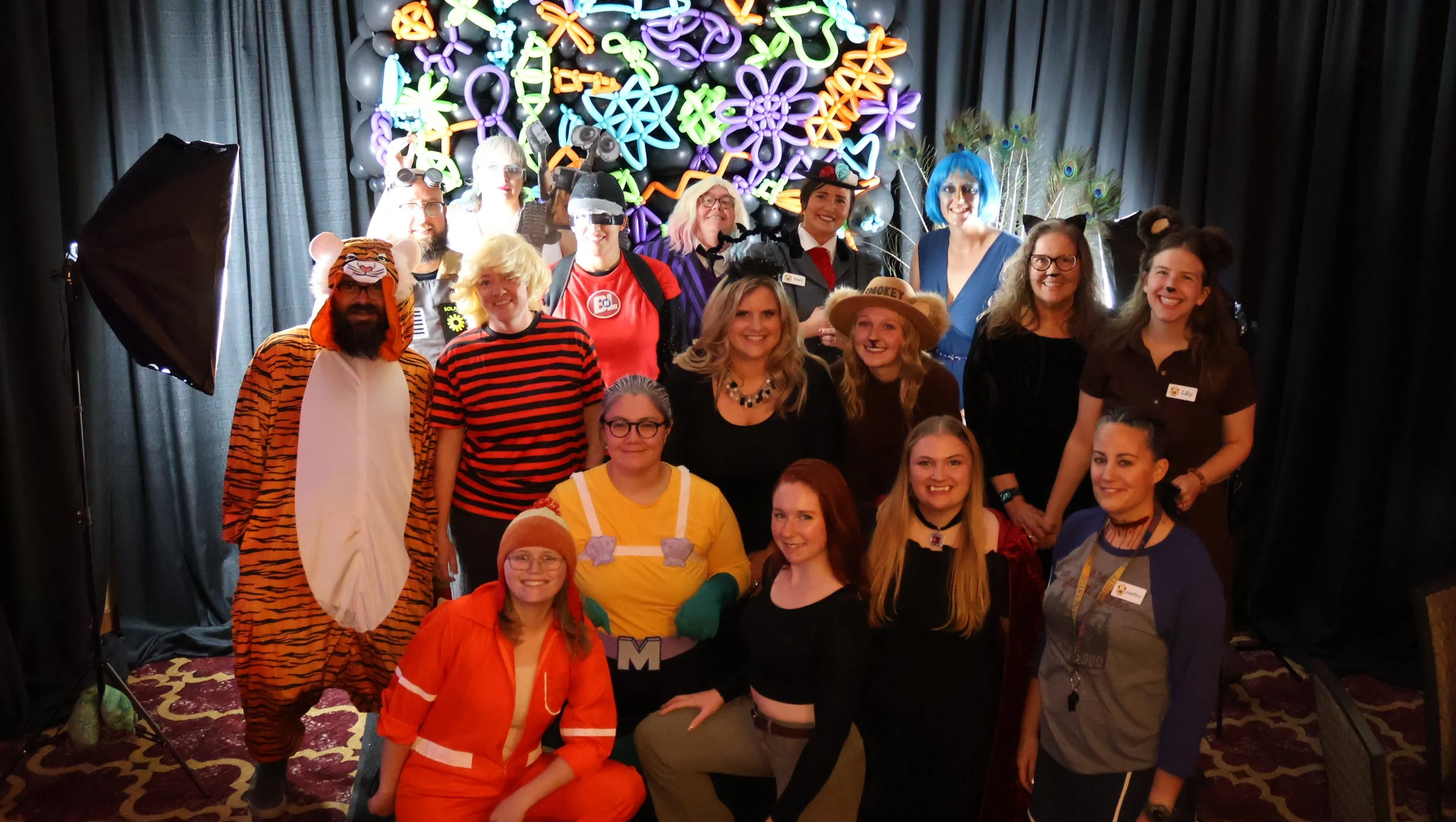 Group of people dressed in colorful costumes and accessories posing for a photo on a stage with a neon light backdrop and black curtains.
