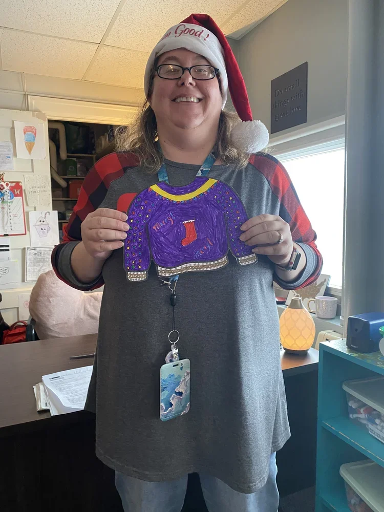 A smiling woman wearing a Santa hat and glasses, holding a colorful drawing of an ugly Christmas sweater with a red stocking. She is in a room with holiday decorations and books, standing near a window.