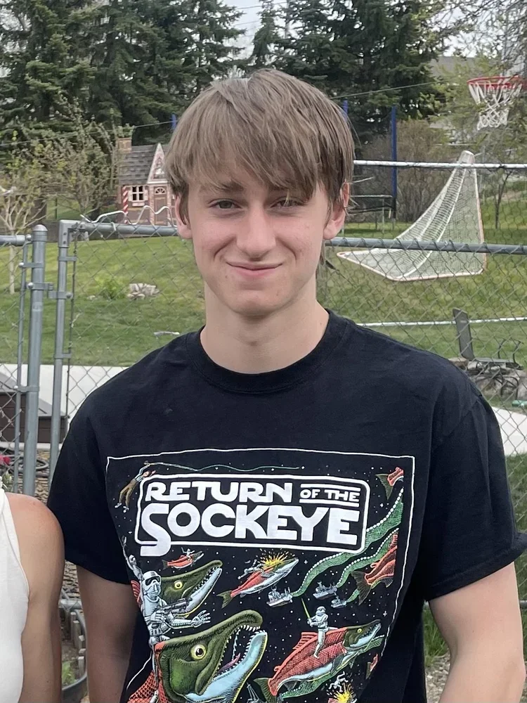 A teenage boy with light brown hair and a slight smile, wearing a black graphic T-shirt featuring dinosaurs and space themes, standing outside in a backyard with a chain-link fence, trees, a small playhouse, and a basketball hoop in the background.