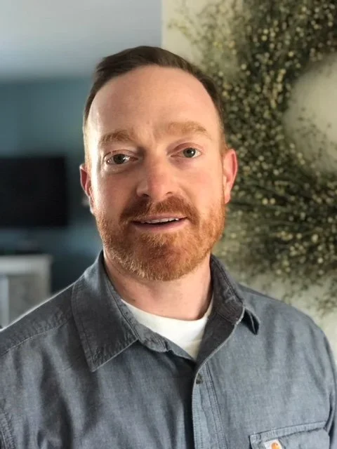A man with short brown hair and a beard, wearing a gray button-up shirt, standing indoors with a blurred background and some greenery or flowers.