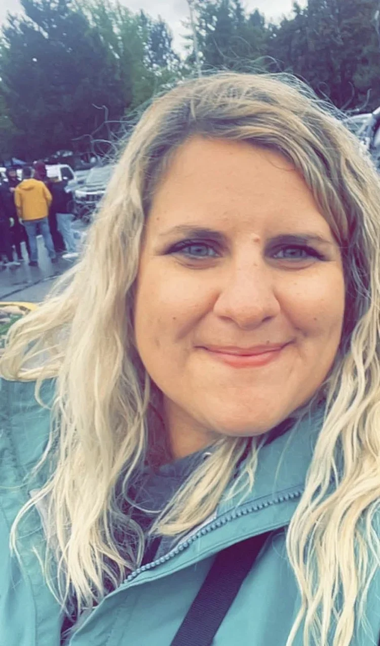 Close-up of a smiling woman with long, wavy blonde hair and blue eyes, wearing a teal jacket in a parking lot with several cars and trees in the background.
