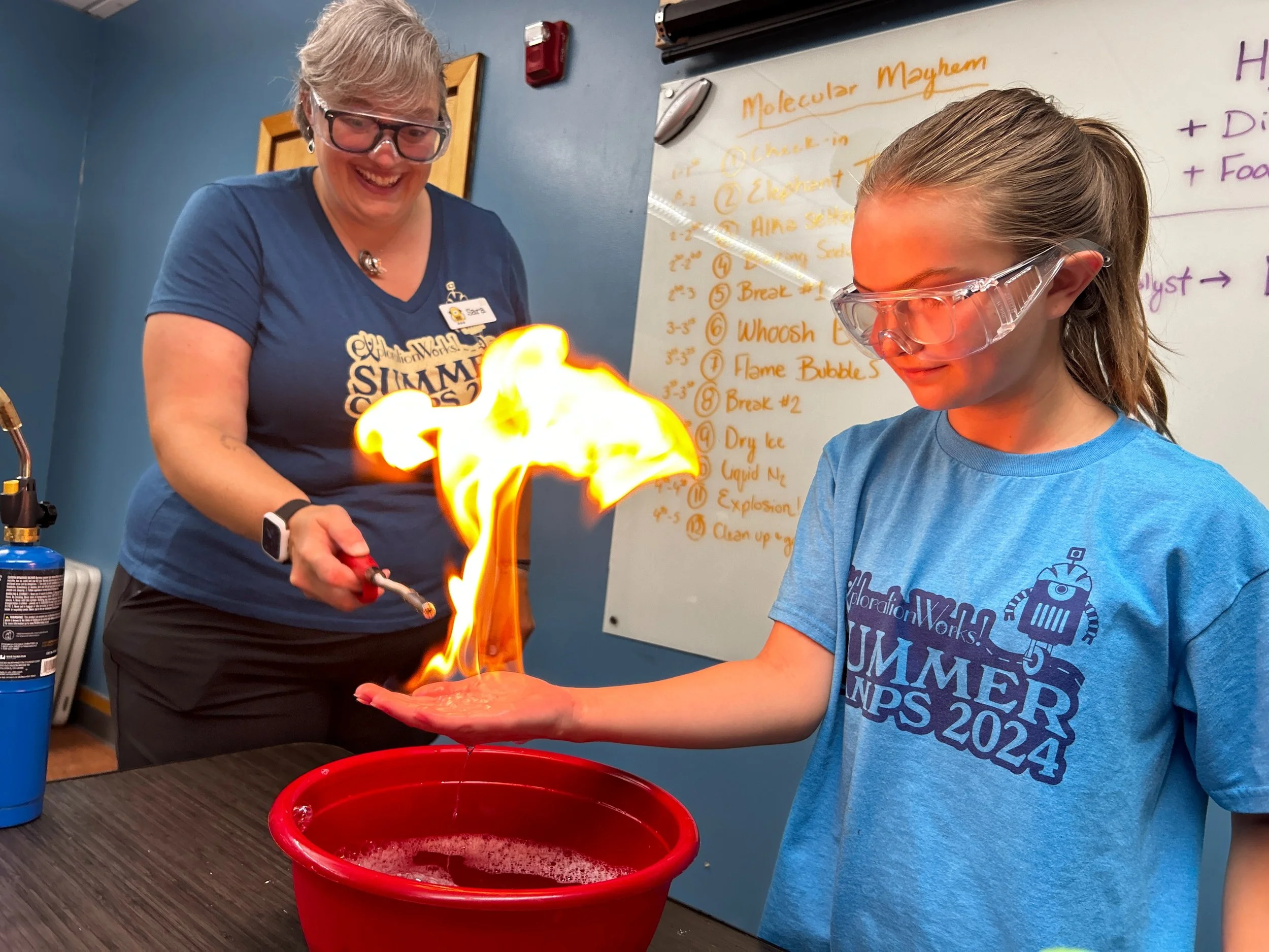 A young girl and an instructor are conducting a science experiment involving fire in a room with a whiteboard and a blue wall. The girl is holding her hand over a red bucket, with flames coming from a substance on her hand. The instructor is smiling and holding a lighter. Both are wearing safety goggles, and the girl is wearing a blue T-shirt with a summer camp logo.