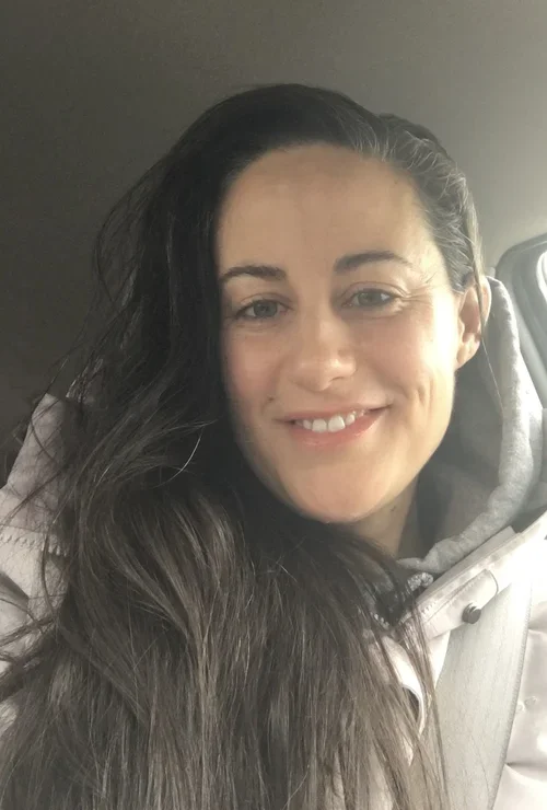 A woman with long dark hair smiling, sitting in a car, wearing a light-colored jacket and a gray hoodie.
