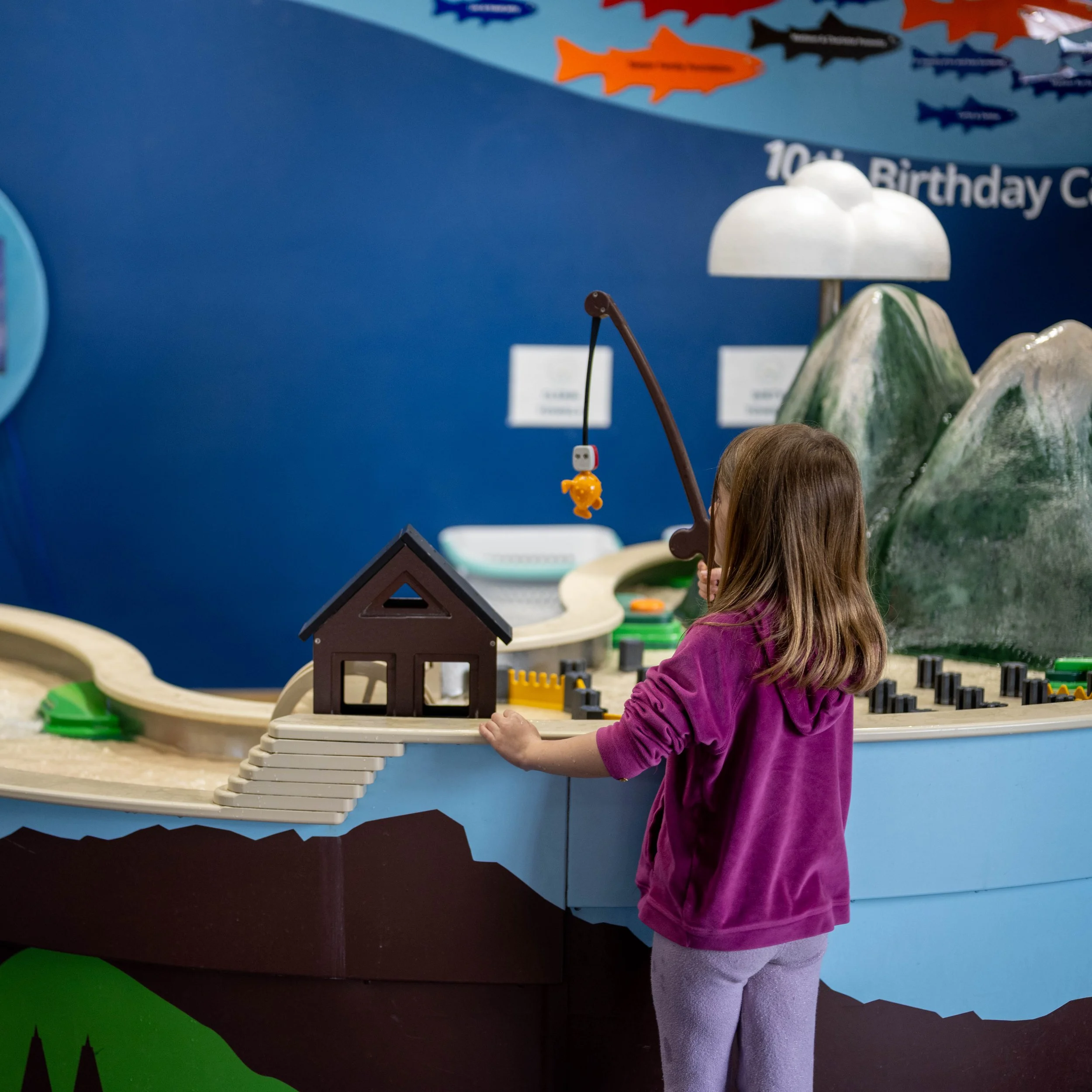 A young girl with long brown hair wearing a purple hoodie and light gray pants playing with a toy train set on a display table at a museum or exhibit. The background features painted mountains, clouds, and a blue wall with fish decorations and a sign reading '10th Birthday'.