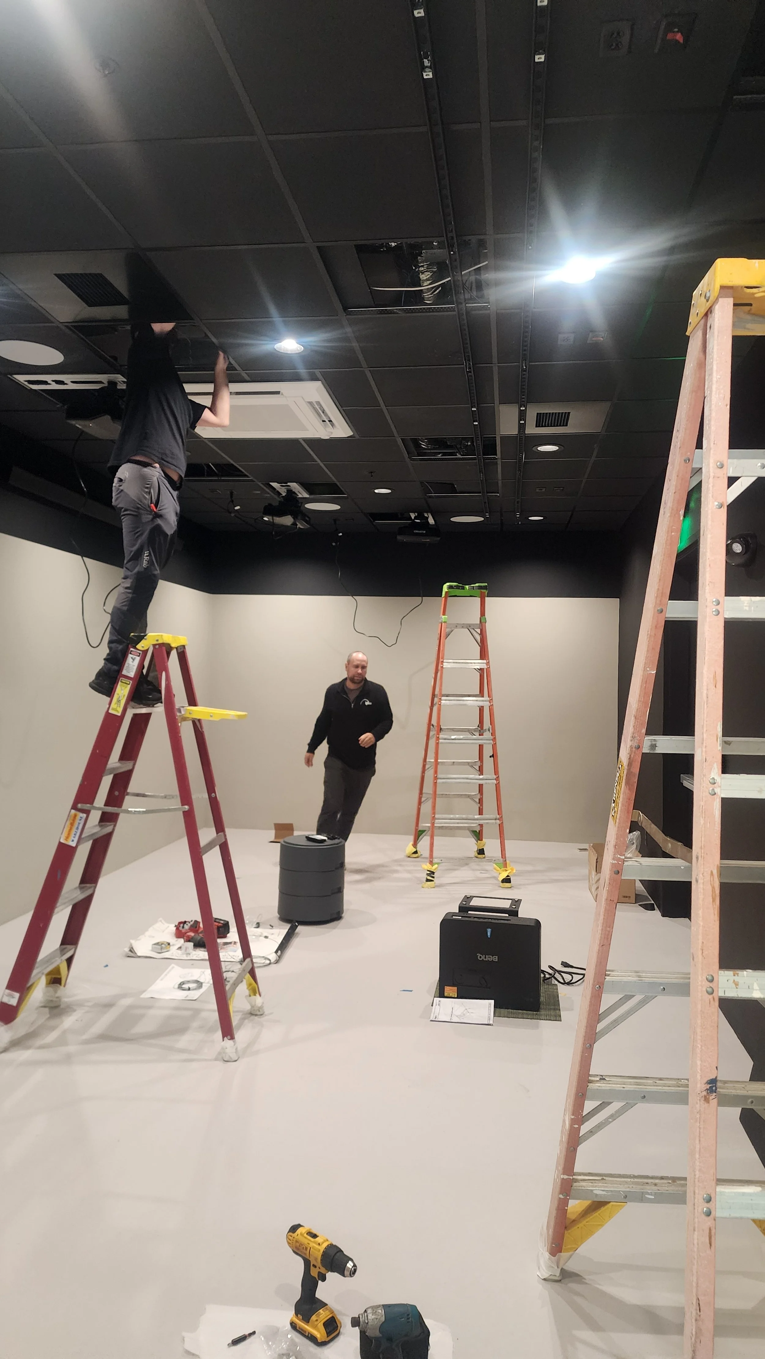 Two workers installing or repairing a ceiling with ladders and tools in an indoor space.