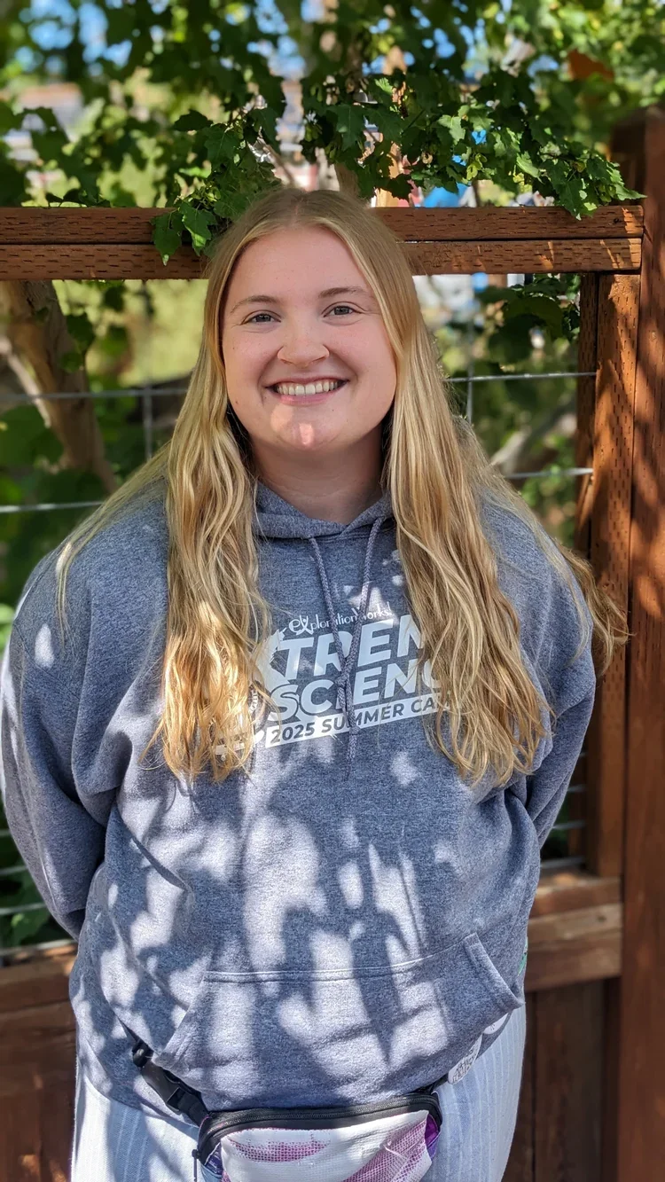 A young woman with long blonde hair smiling outdoors in front of a wooden fence and leafy tree, wearing a gray hoodie with white text and a small waist pack.