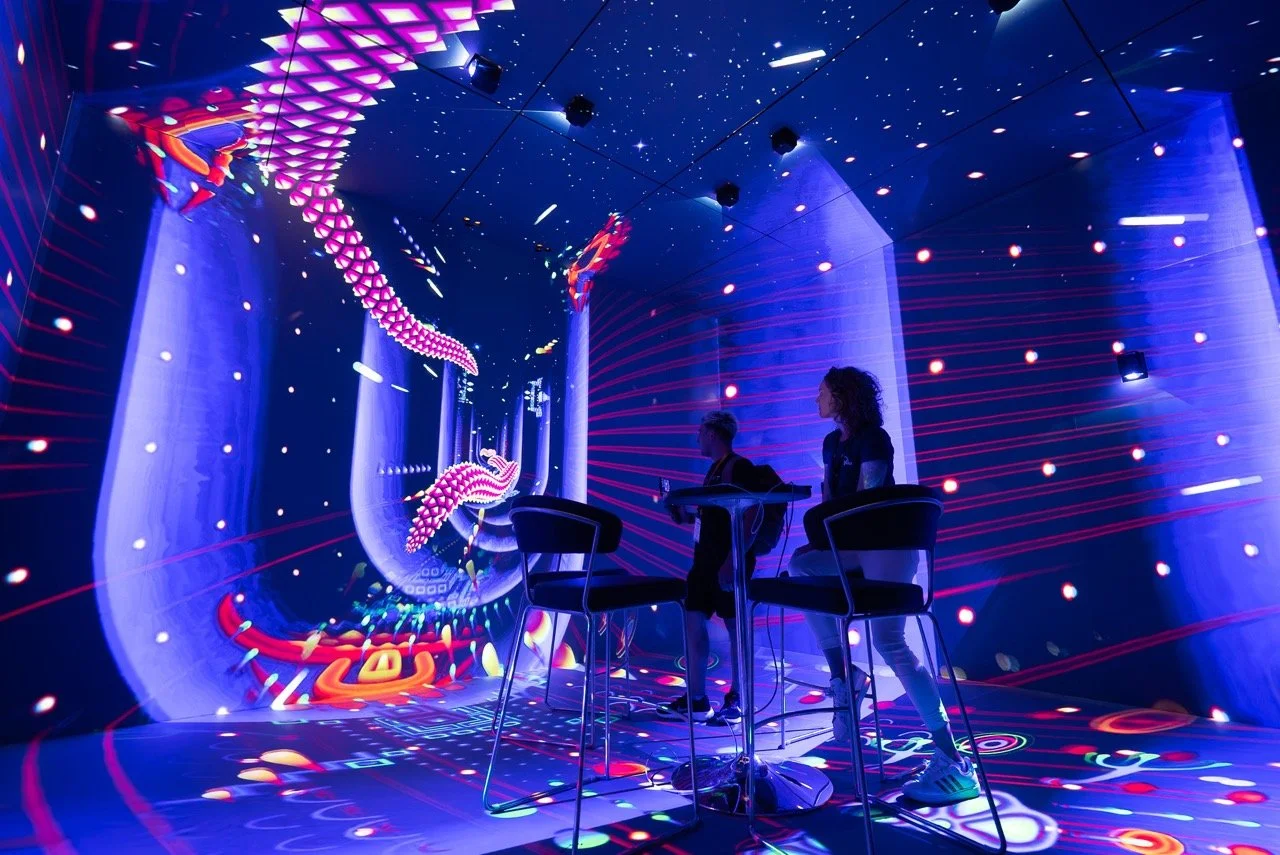 Two DJ performers are in a neon-lit, virtual reality-themed room with colorful, glowing digital effects and a large projection of a swirling dragon on the wall.