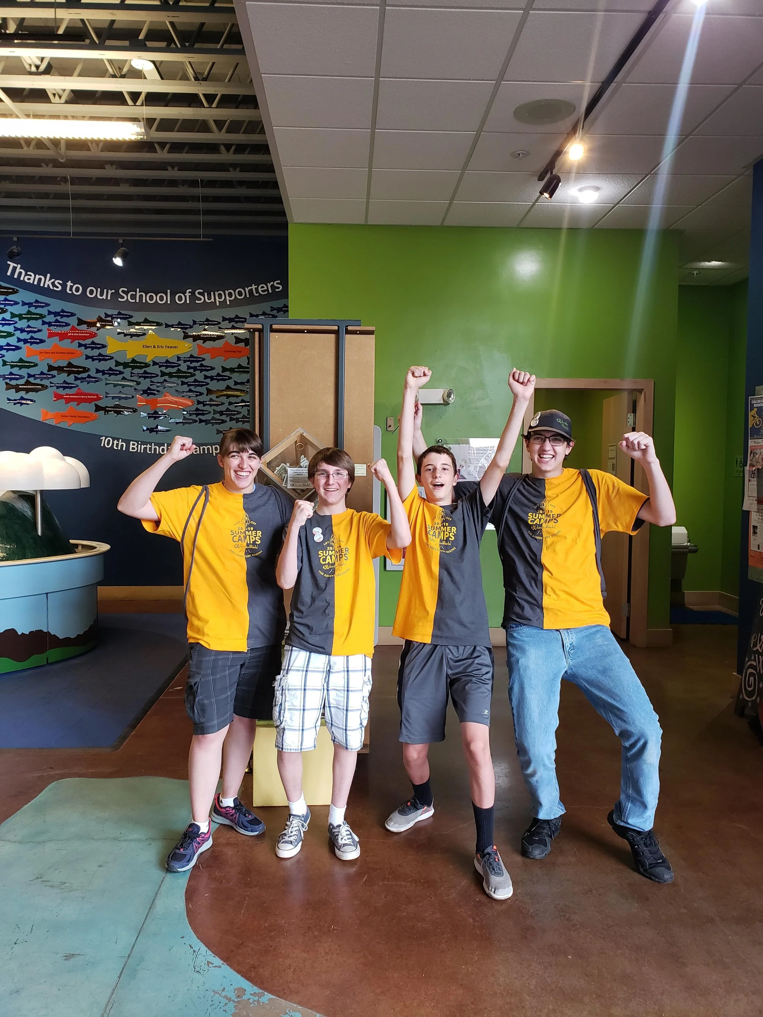 Four teens celebrate with their arms raised inside a colorful indoor space, wearing yellow and gray t-shirts for a summer camp event.