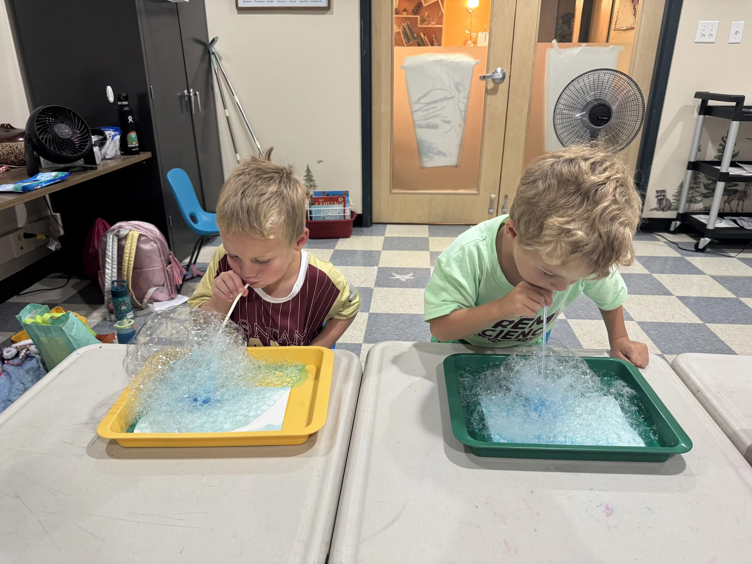 Two young boys blowing bubbles into trays of soapy water in a classroom setting.