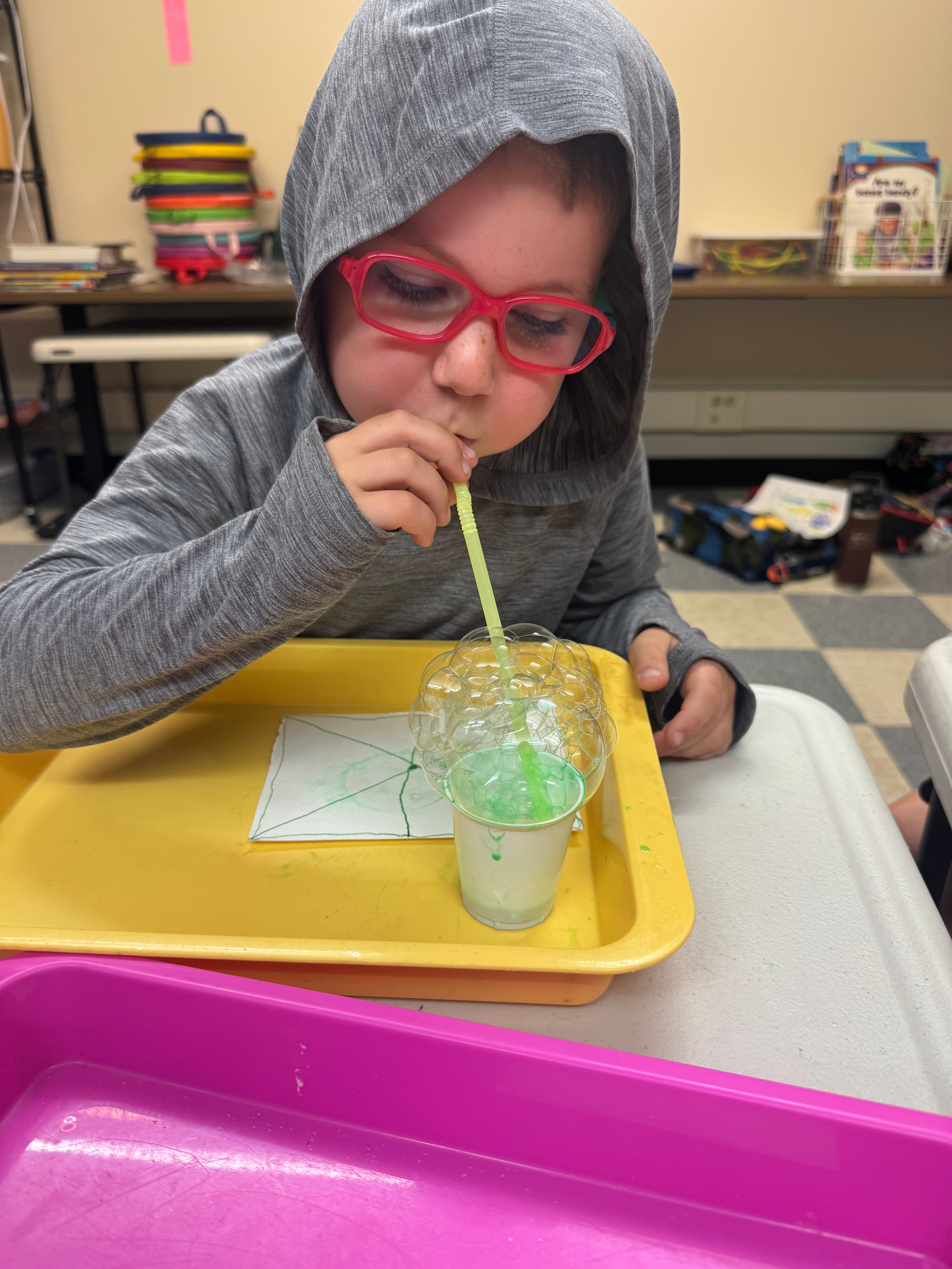 A boy wearing a gray hoodie and red glasses is blowing bubbles through a straw into a cup of green soap solution.