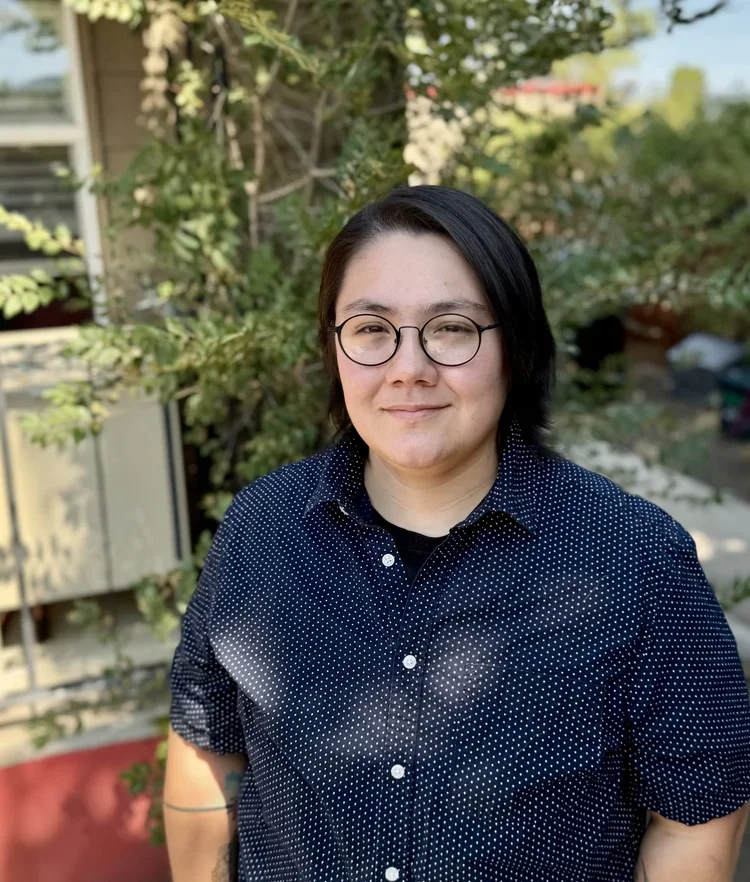 A person with black hair, glasses, and a black polka-dot shirt standing outdoors a garden or backyard with greenery and flowers.