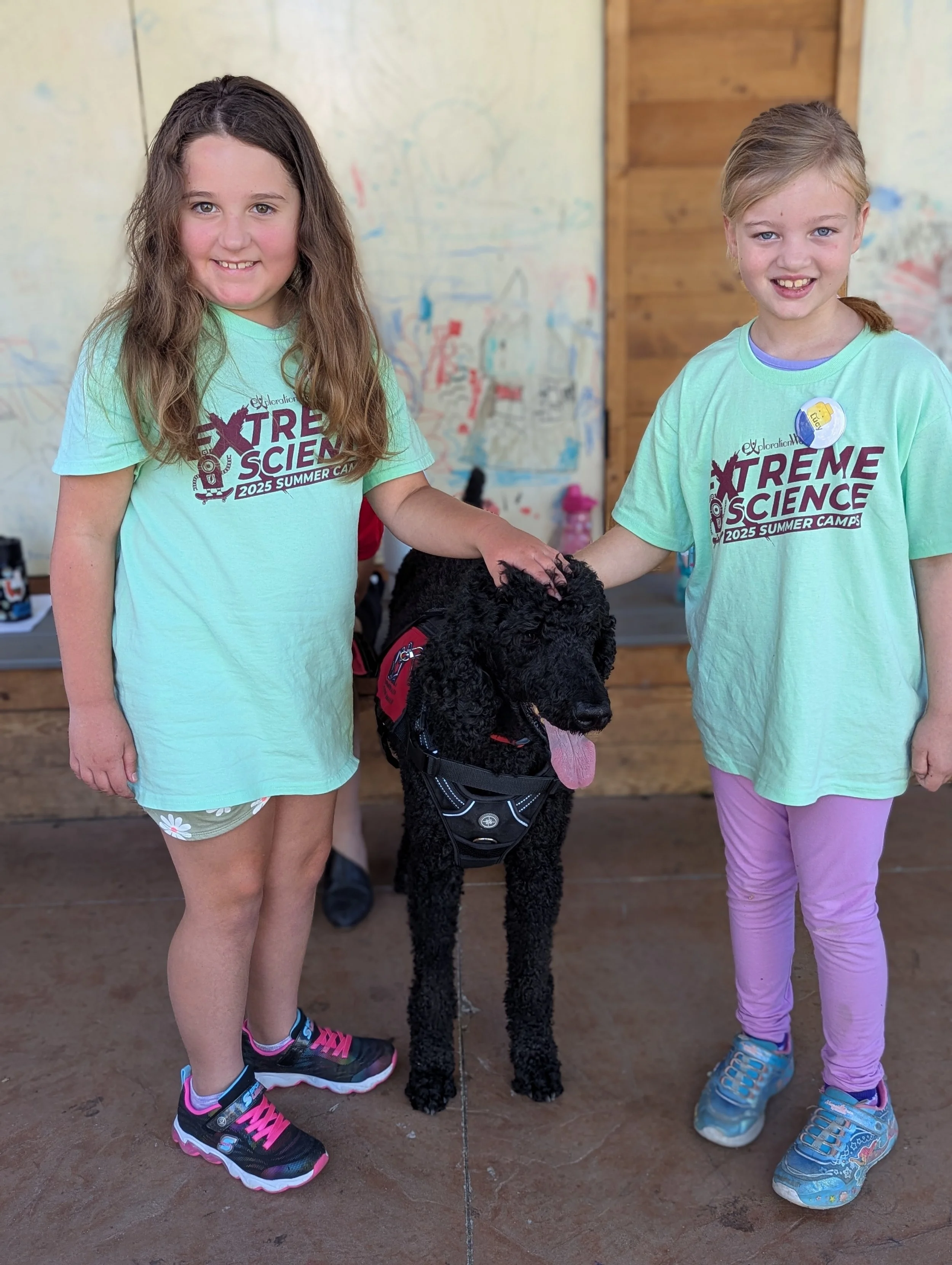 Two young girls smiling at the camera, petting a black standard poodle dog, in a room with a wooden wall and colorful drawings on the wall behind them. They are wearing light green T-shirts with 'Extreme Science Summer Camp 2025' printed on them.