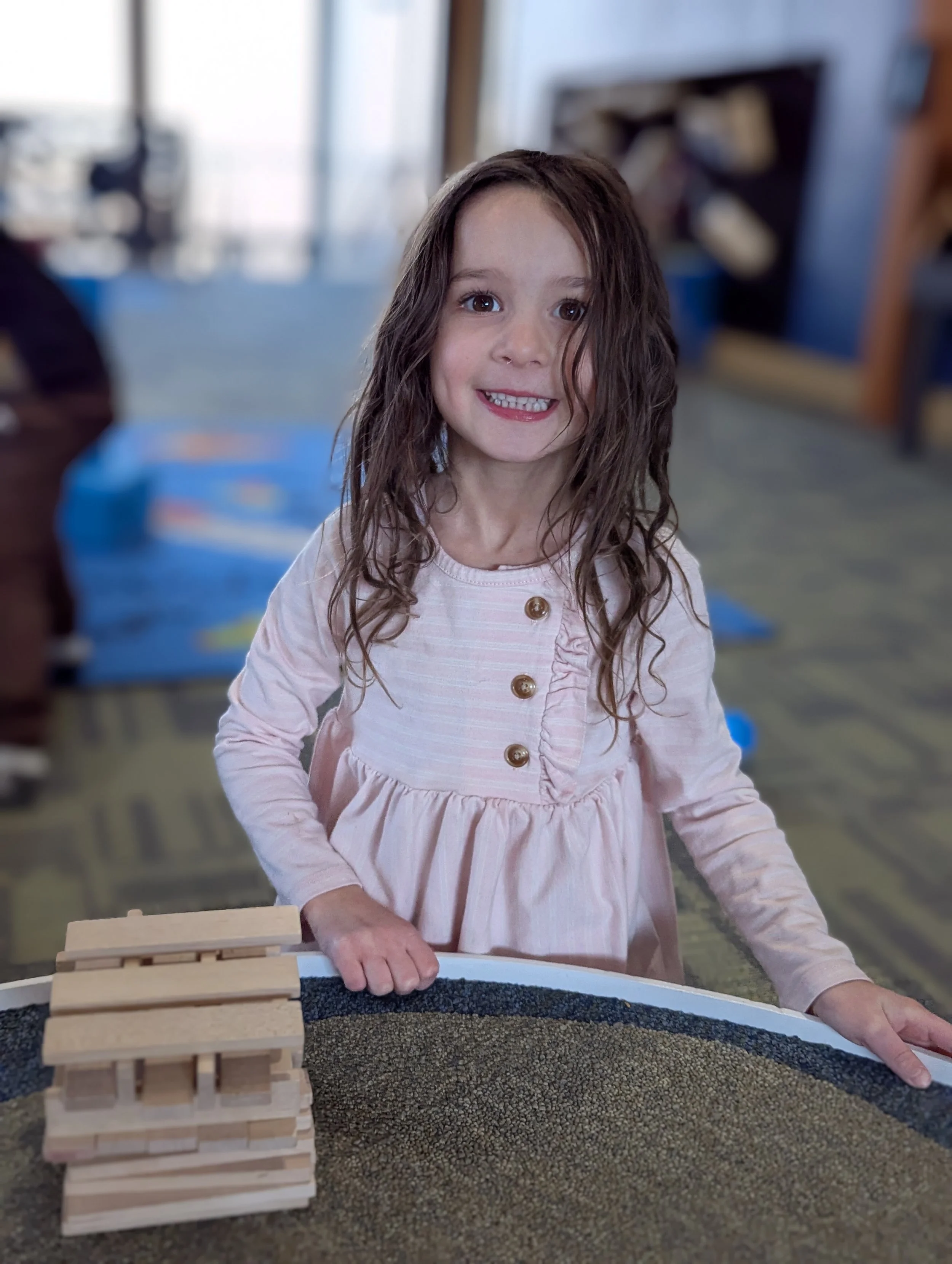 A young girl with long, curly brown hair and a big smile, wearing a light pink dress with gold buttons, standing at a table with a stack of wooden blocks.