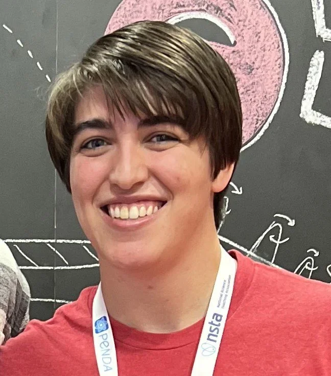 A young man with brown hair, smiling, wearing a red shirt and a lanyard, standing in front of a chalkboard with drawings and equations.