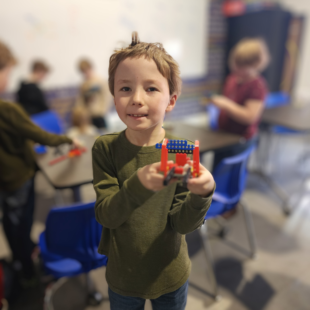 A young boy smiling and holding a LEGO creation in a classroom with other children working at tables in the background.