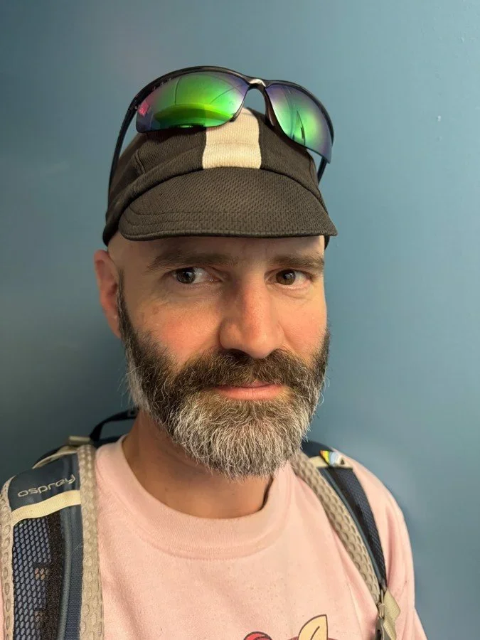 A man with a beard and mustache, wearing a brown cap and sunglasses on top, pink t-shirt, and a backpack, standing against a blue wall.