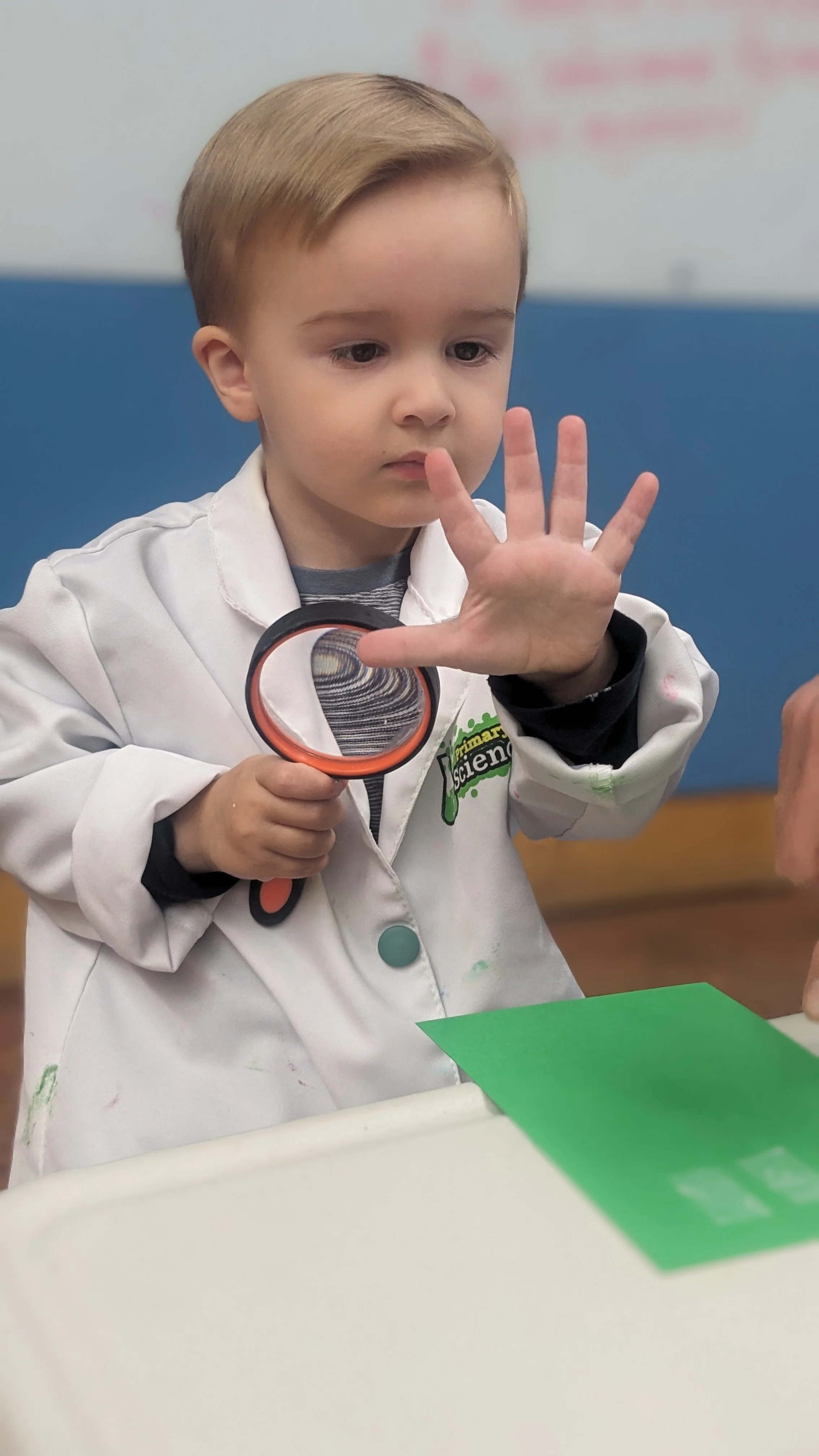 Young boy in a science lab coat holding a magnifying glass and examining his hand with a focused expression.