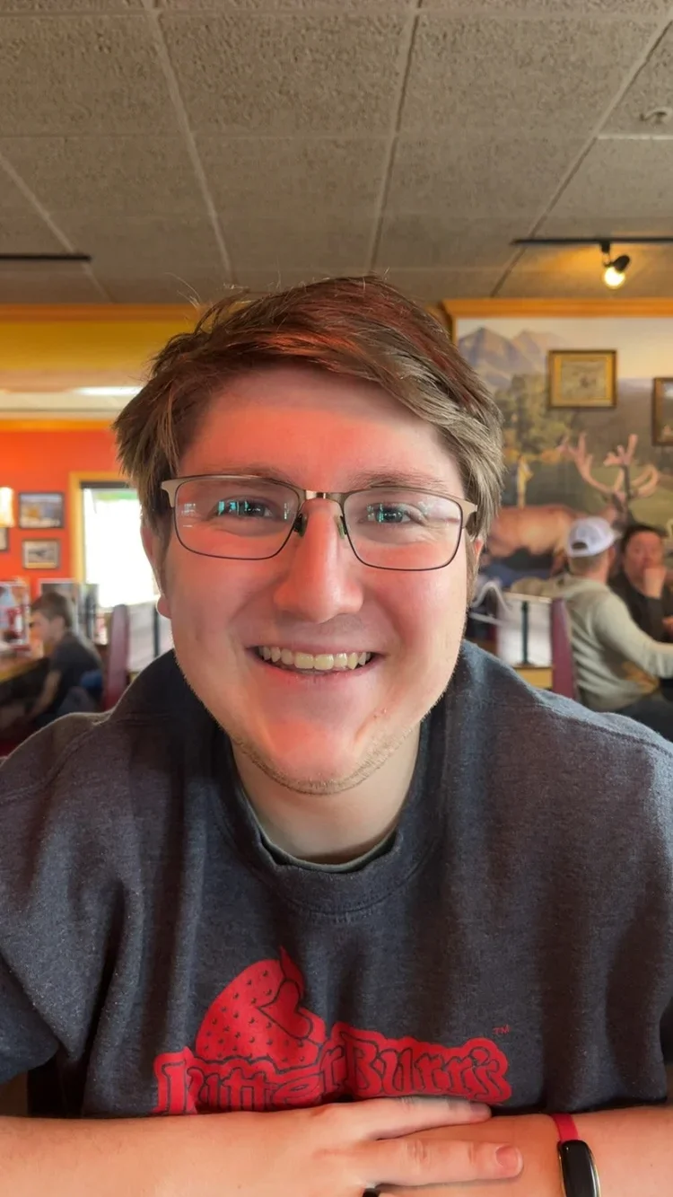 A young man with glasses smiling at a restaurant, with other patrons seated at tables and wall decorations including paintings and a mountain scenery in the background.