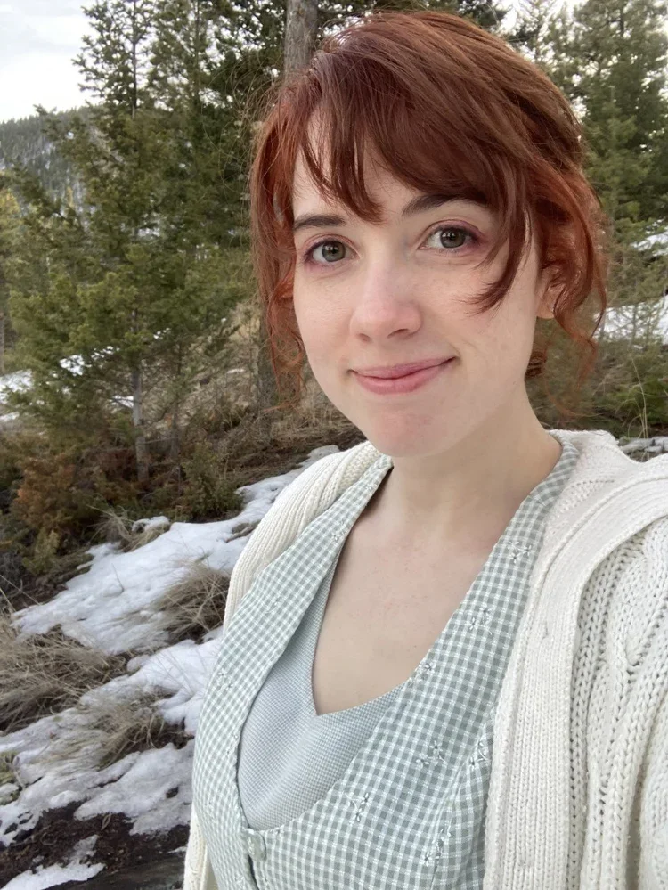 A young woman with short red hair smiling outdoors near a snowy landscape and evergreen trees.