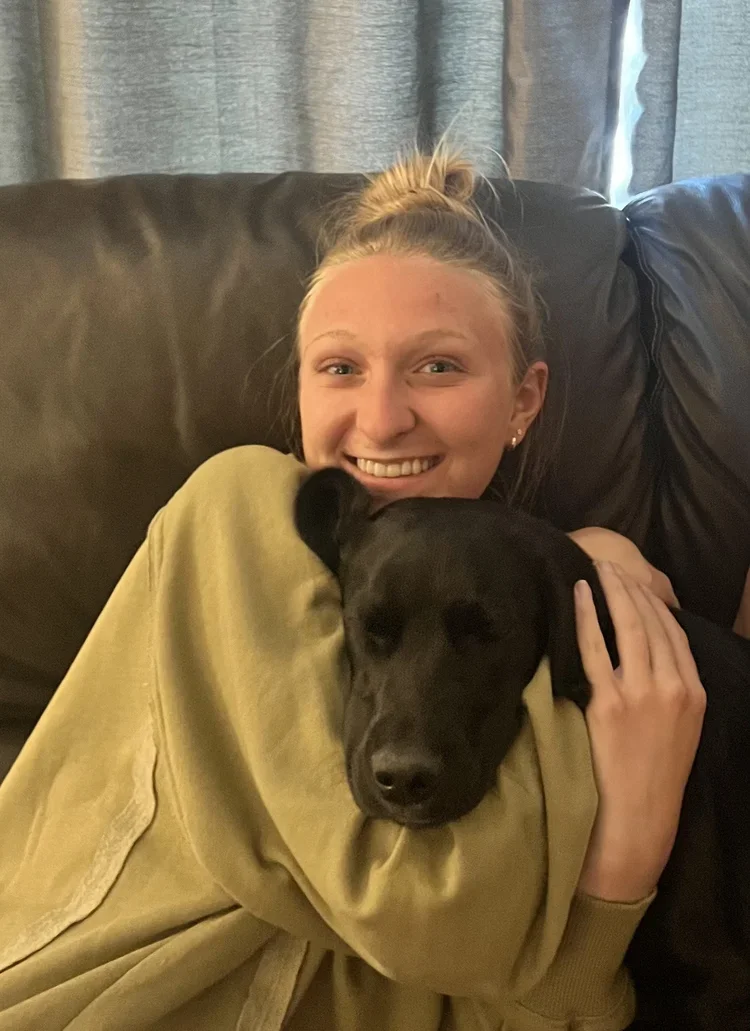 A young woman with blonde hair tied in a bun smiling and hugging a black dog on a couch.
