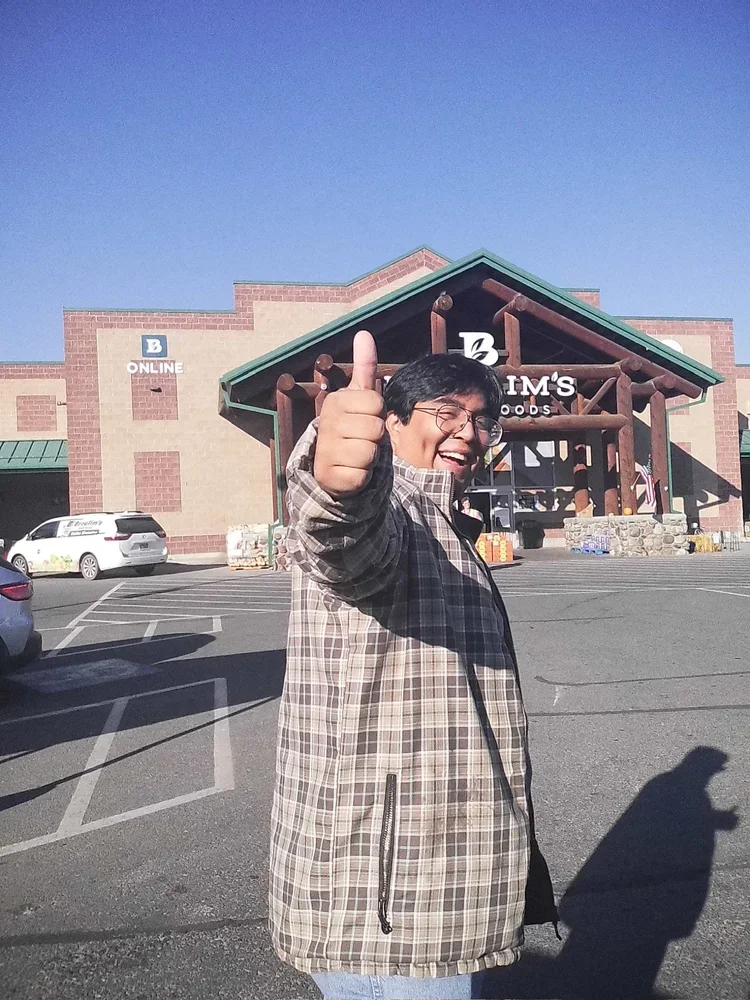 Smiling man wearing glasses and a plaid jacket giving a thumbs-up in front of a grocery store at a parking lot on a sunny day.
