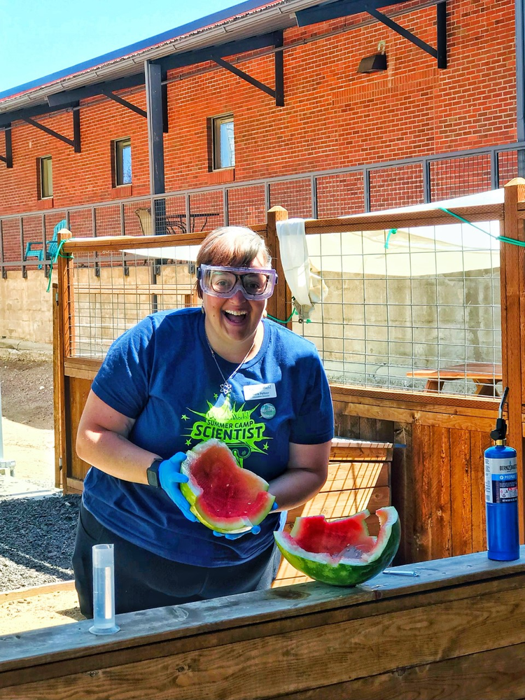 A woman at summer camp summer camp smiling and holding a slice of watermelon outside near a wooden fence and brick building.
