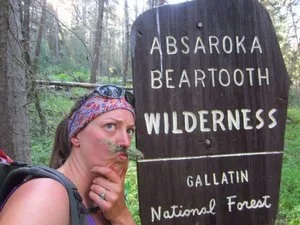 Woman with a bandana taking a selfie next to a wooden sign for Absaroka Beartooth Wilderness in Gallatin National Forest, surrounded by trees.