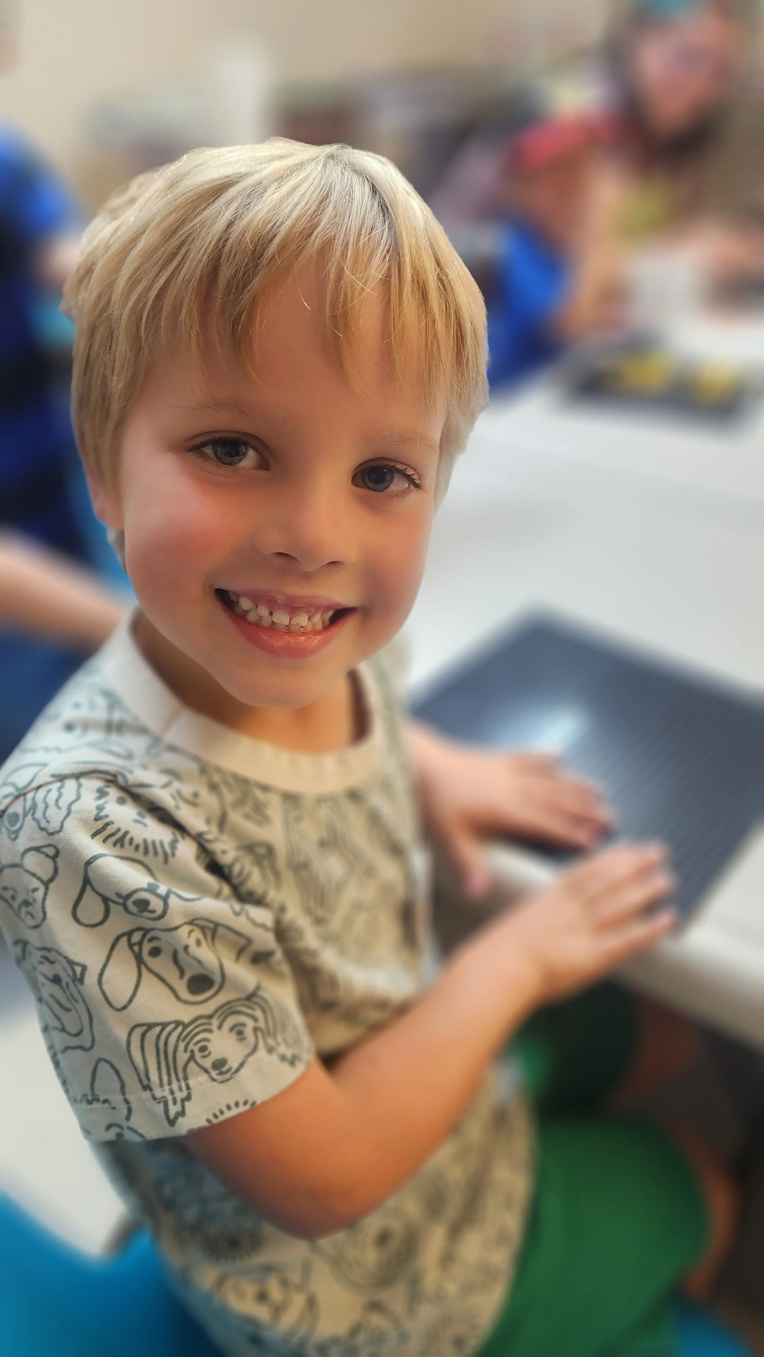 A young boy with blonde hair and blue eyes smiling while using a laptop in a classroom.