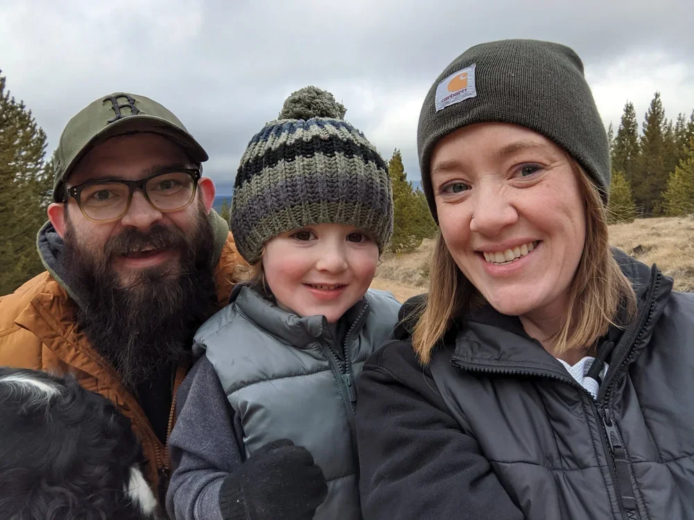 Three people outdoors in a landscape with trees, wearing winter clothing and smiling at the camera. There is a man with glasses and a cap, a woman with a beanie, and a young boy with a knit hat.