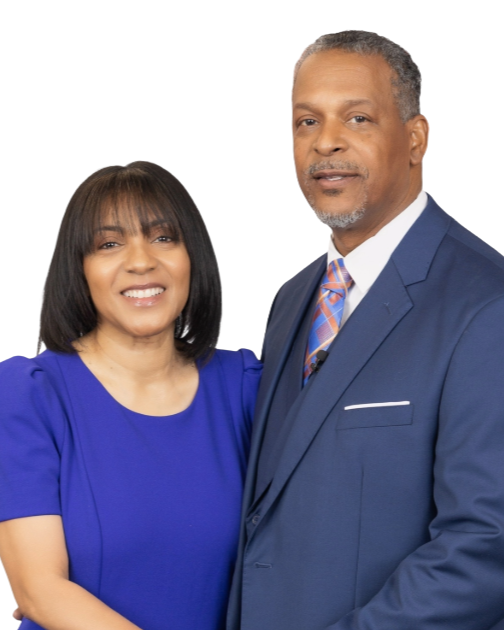 A smiling woman with shoulder-length dark hair in a blue dress standing next to a serious-looking man in a dark blue suit and colorful tie, both facing forward against a white background.