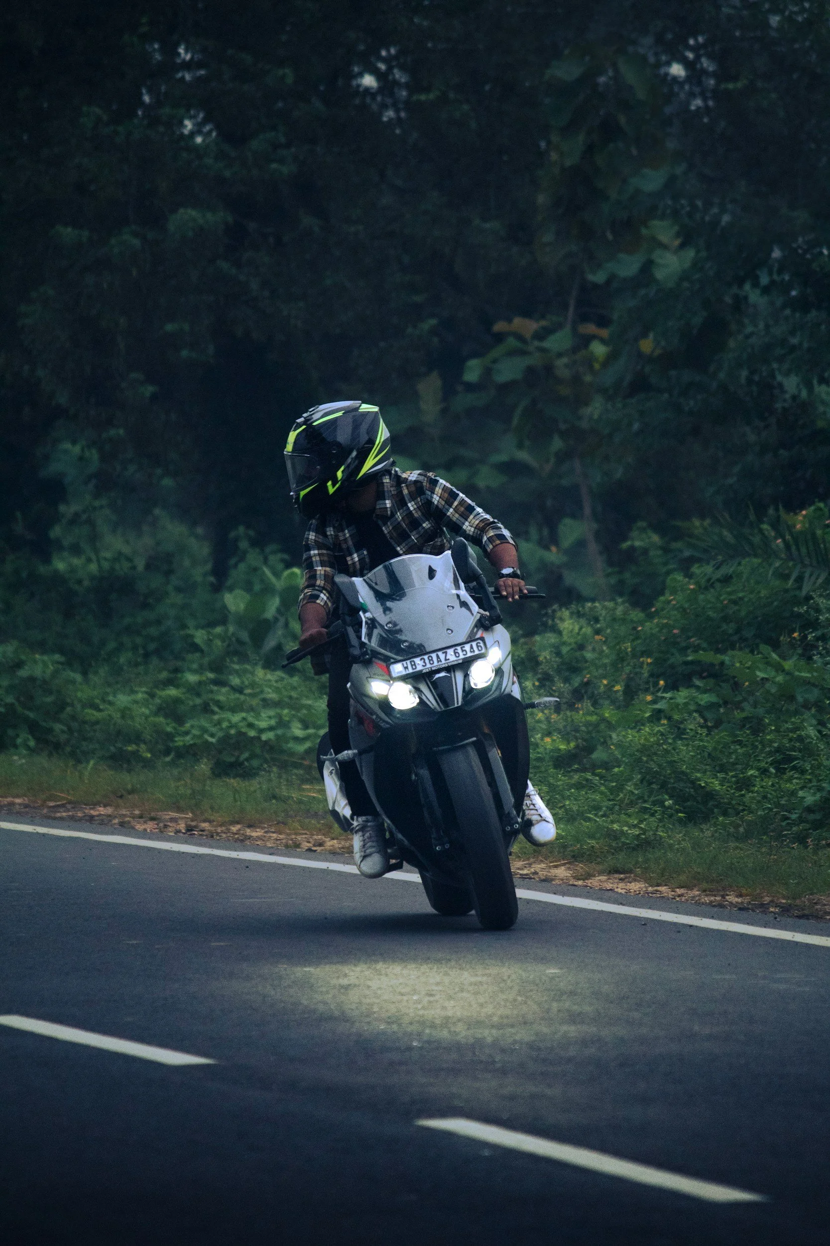 A person riding a motorcycle on a road surrounded by lush green trees, wearing a black helmet, plaid shirt, and white sneakers.