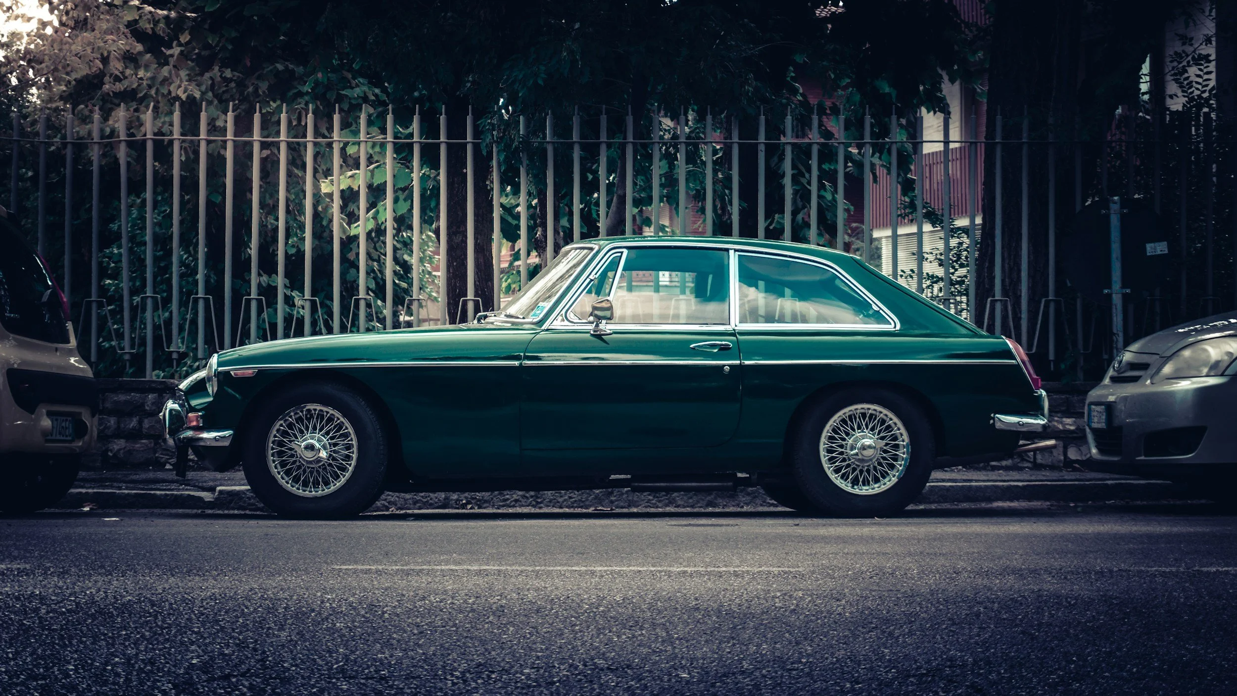 A vintage green car parked between modern vehicles along a city street, with trees and a fence in the background.