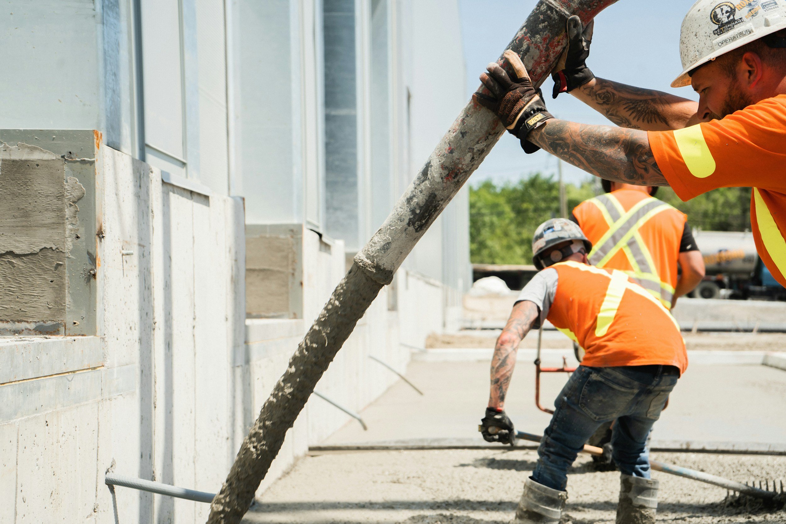 Construction workers in orange safety vests and helmets pouring concrete with a large hose at a construction site.