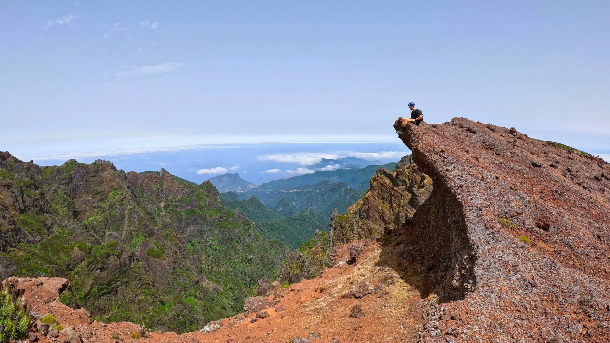 A person wearing a helmet and backpack sitting on a large, flat rock formation high in the mountains.