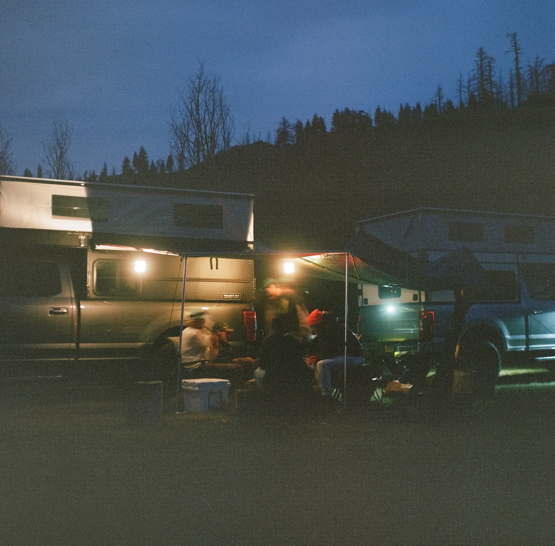 Nighttime scene with two parked camper vans, illuminated by lights, and a group of people sitting under a canopy between the vans, with a hill and bare trees in the background.