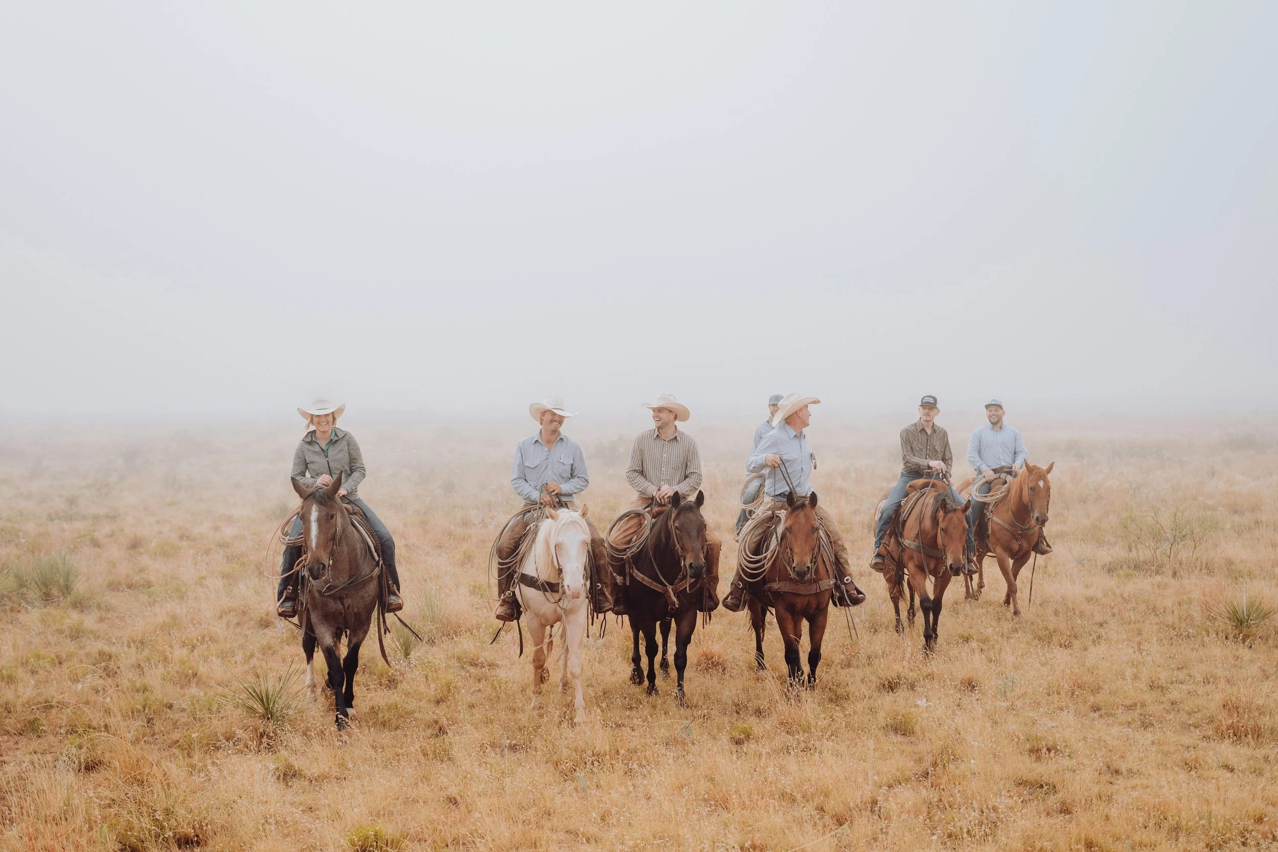 A group of six people riding horses through a foggy, open grassland landscape, all wearing hats.