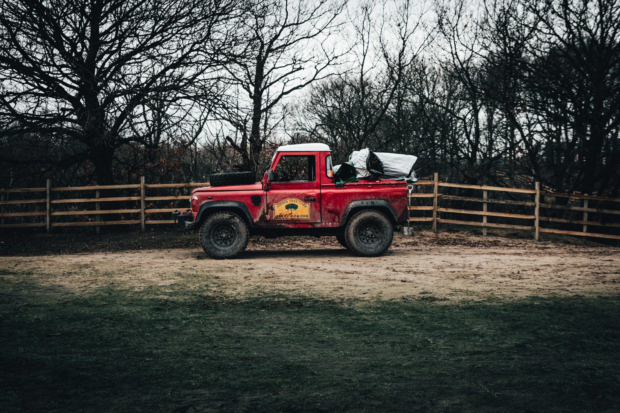 Red pickup truck parked behind a wooden fence with trees in the background, overcast sky.
