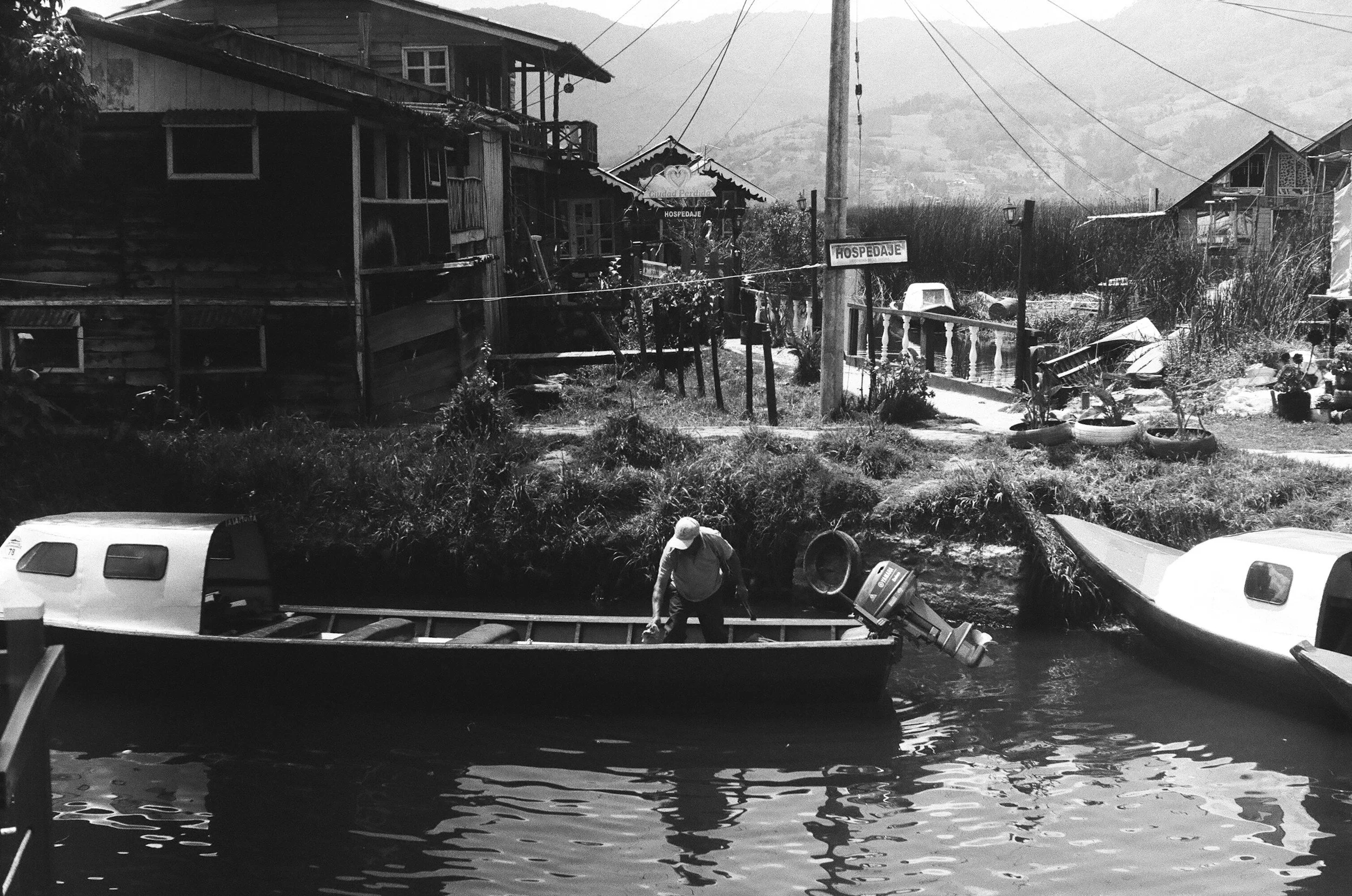 A man in a boat on a river with houses and boats on the riverbank in the background.