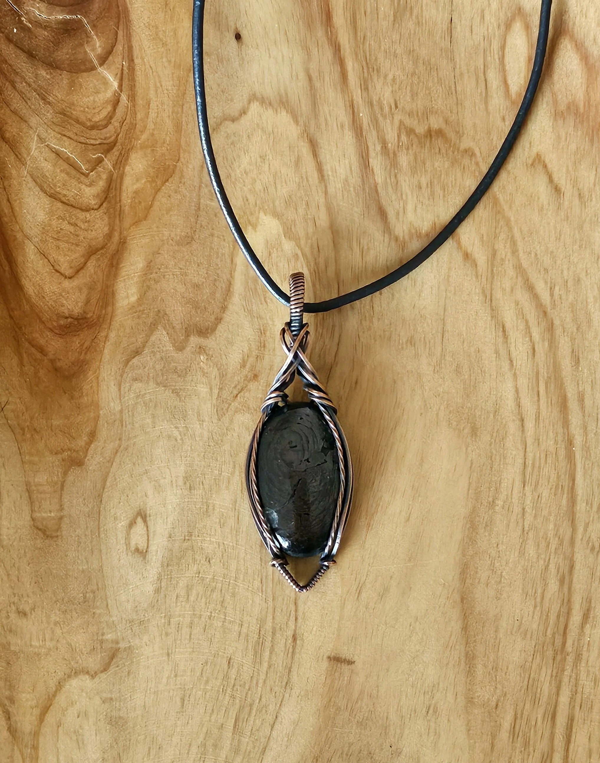 A handcrafted pendant necklace with a dark, oval-shaped stone wrapped in wire, hanging from a black cord.