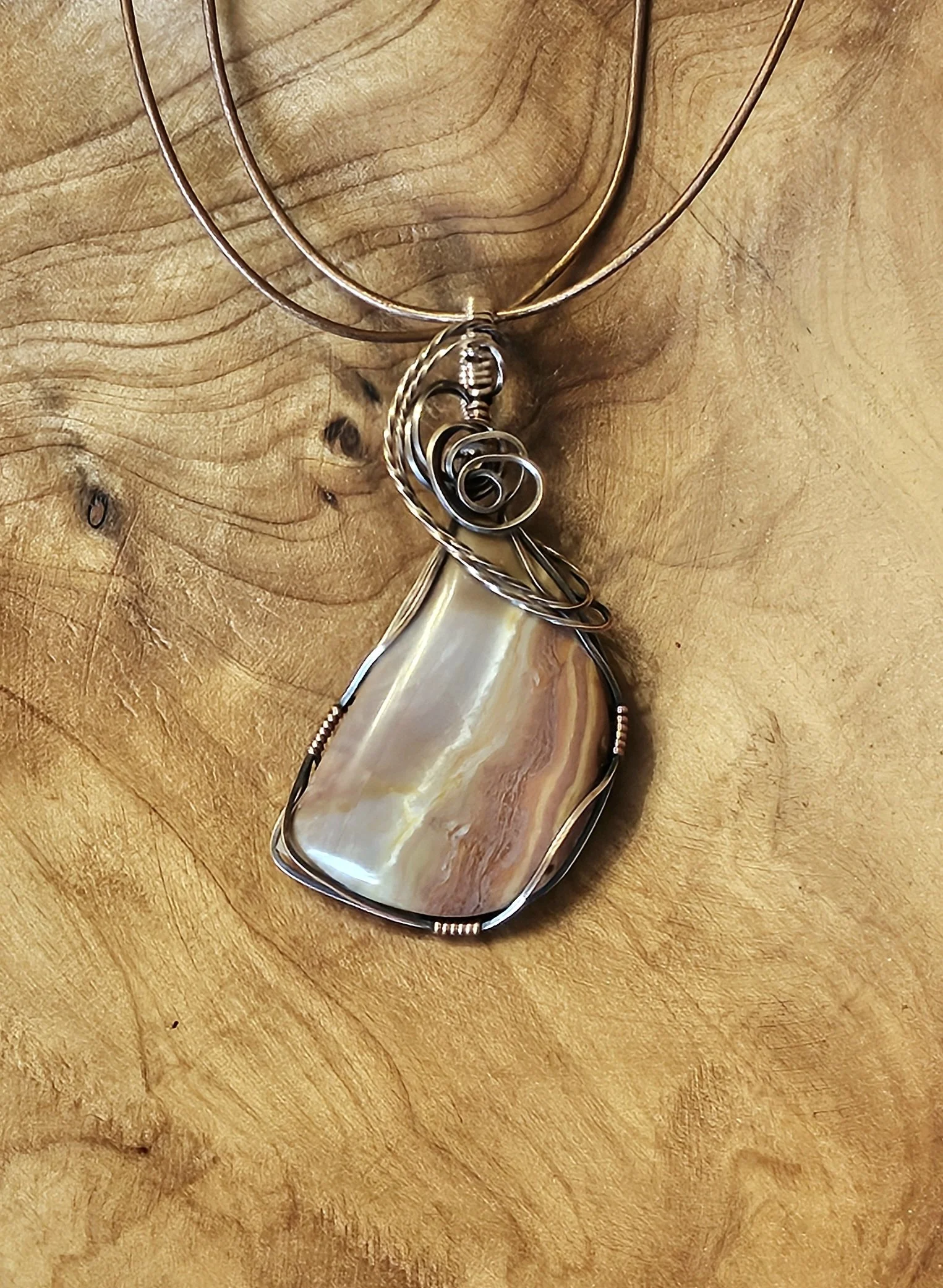 A pendant necklace featuring a large, irregularly shaped seashell attached to a wire-wrapped metal frame, resting on a wooden surface.