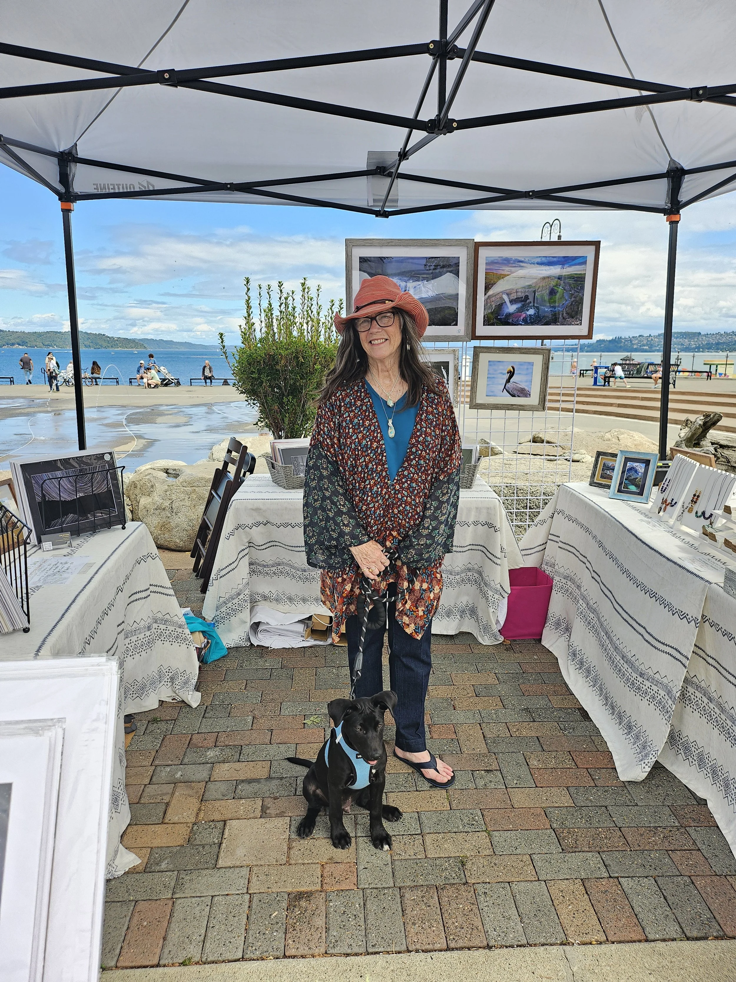 Woman standing with a small black dog in front of outdoor art booth with paintings and photographs, near a waterfront with people and a cloudy sky.