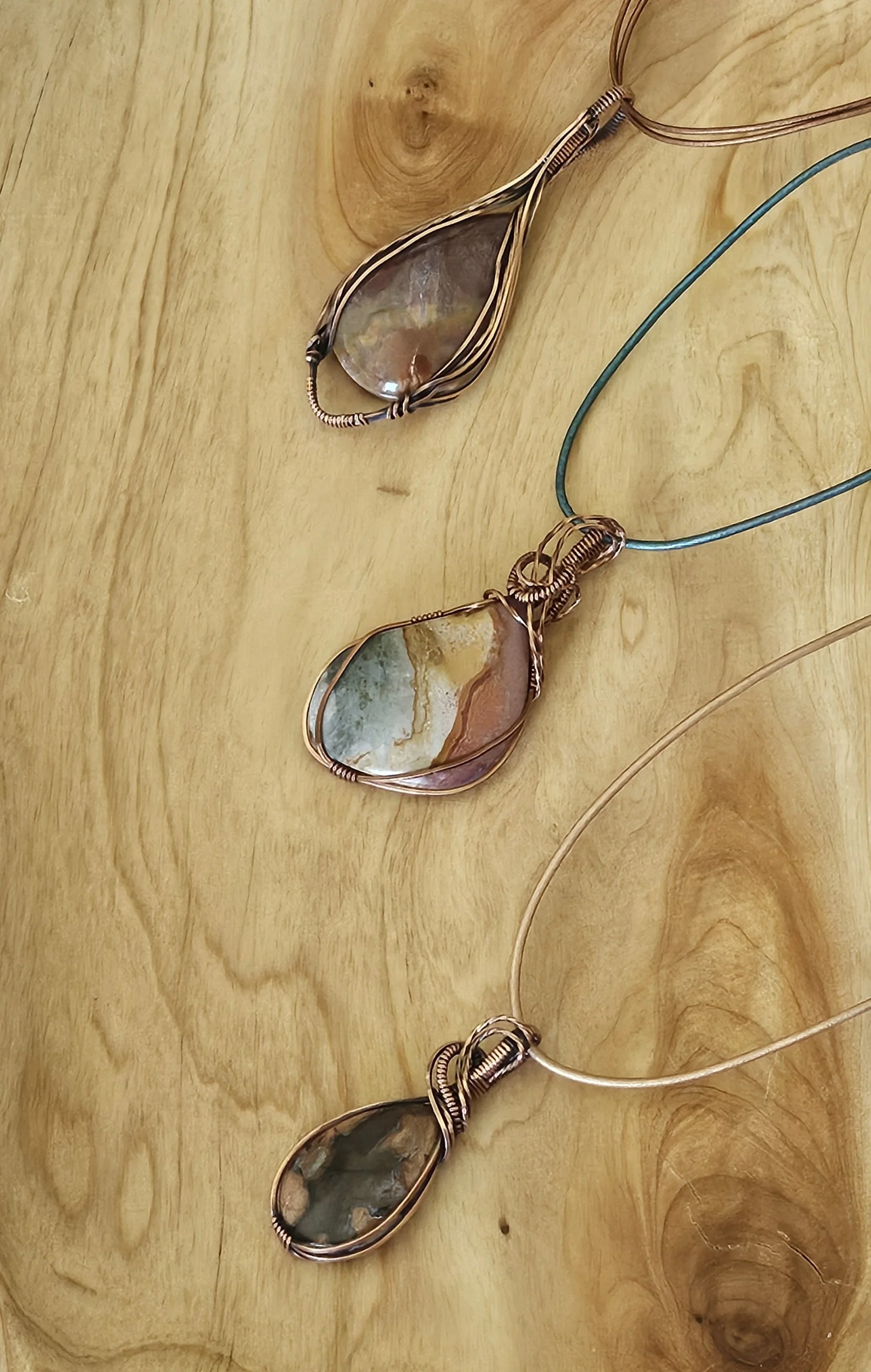 Three jewelry necklaces with large, multicolored stones wrapped in intricate wire designs, laid on a wooden surface.