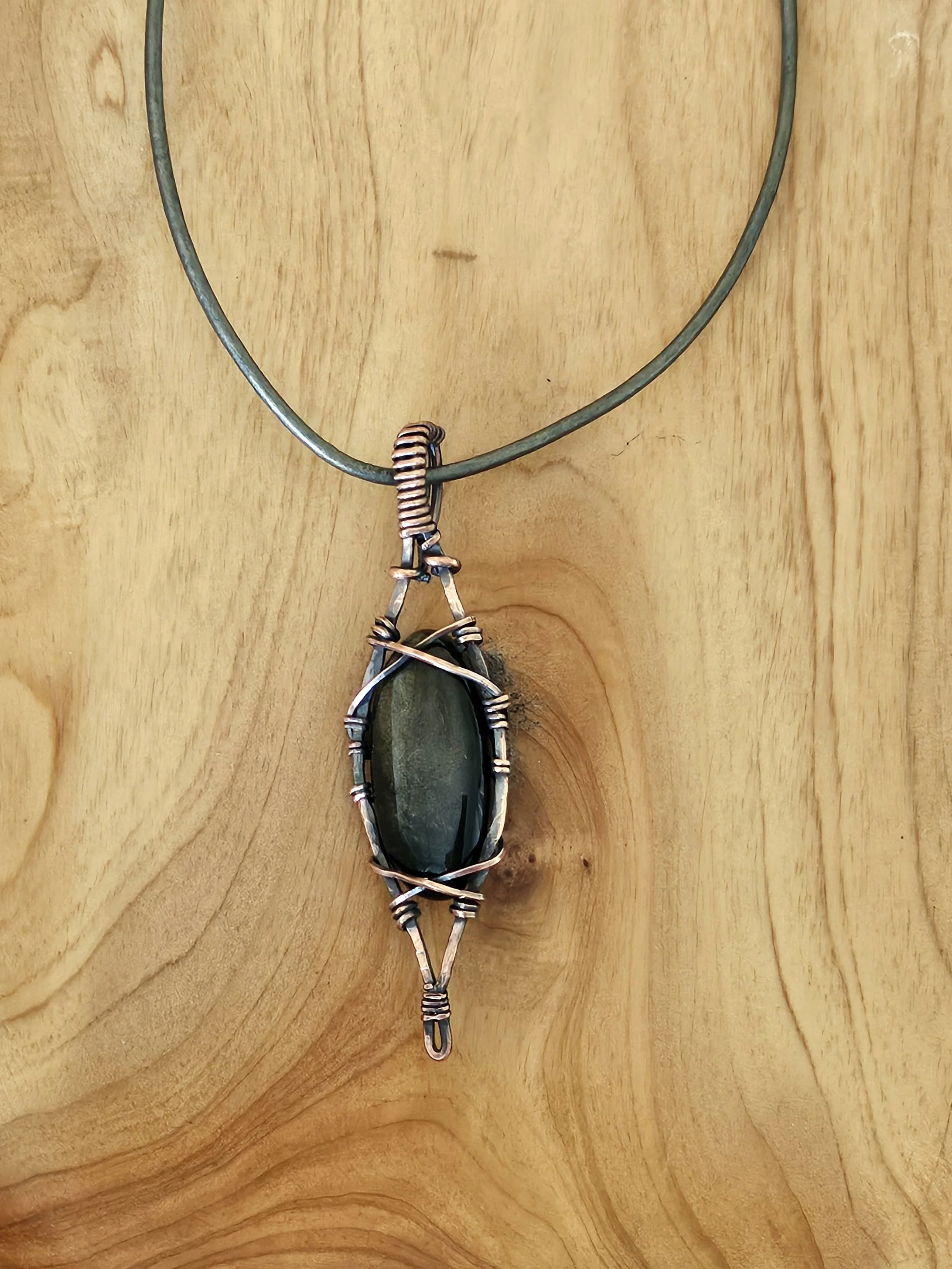 Handmade wire-wrapped pendant with a dark green gemstone on a metal cord necklace displayed against a wooden background.