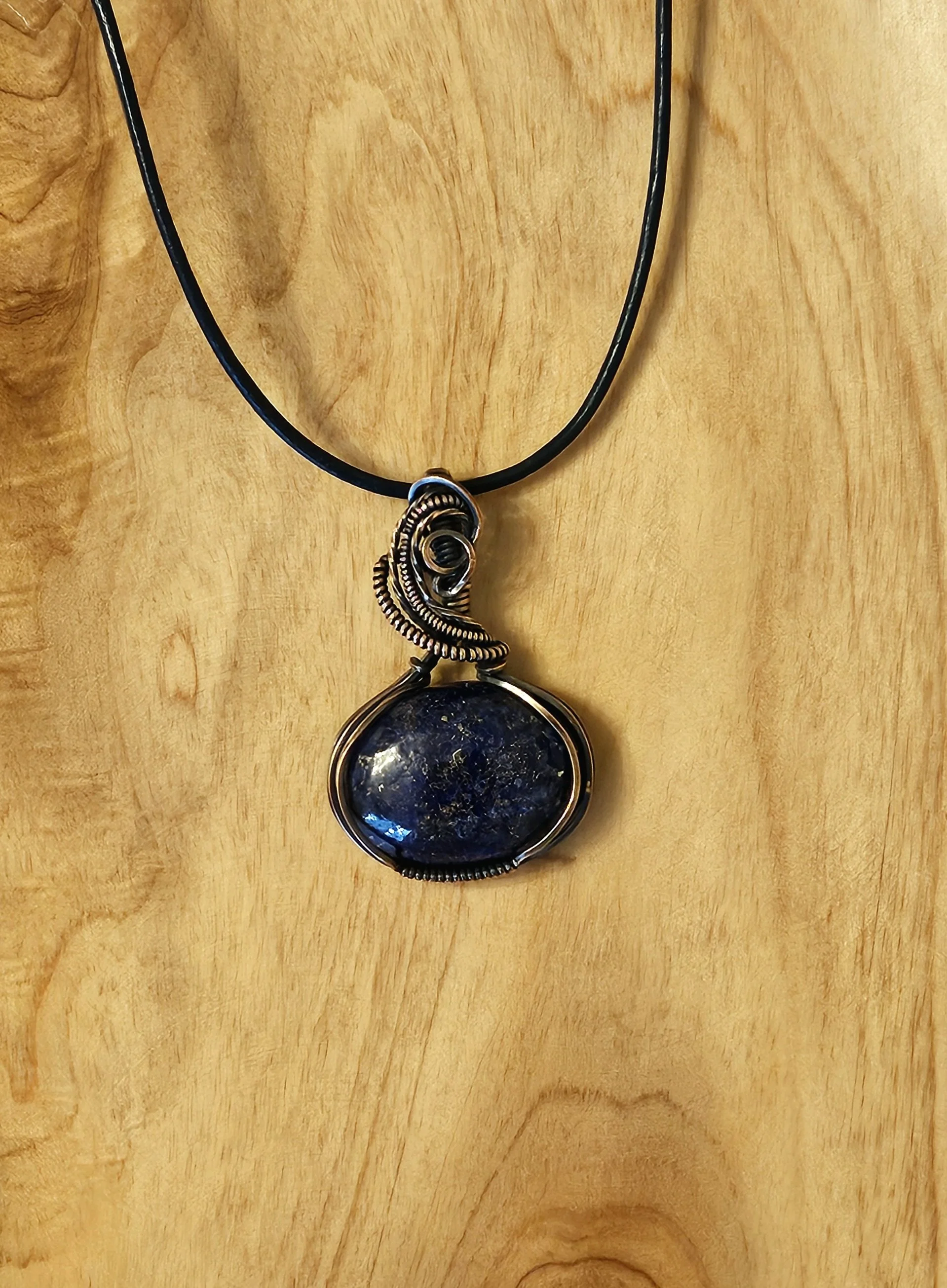 A pendant necklace with a dark, round stone set in a wire-wrapped metal frame, hanging from a black cord, displayed on a wooden surface.