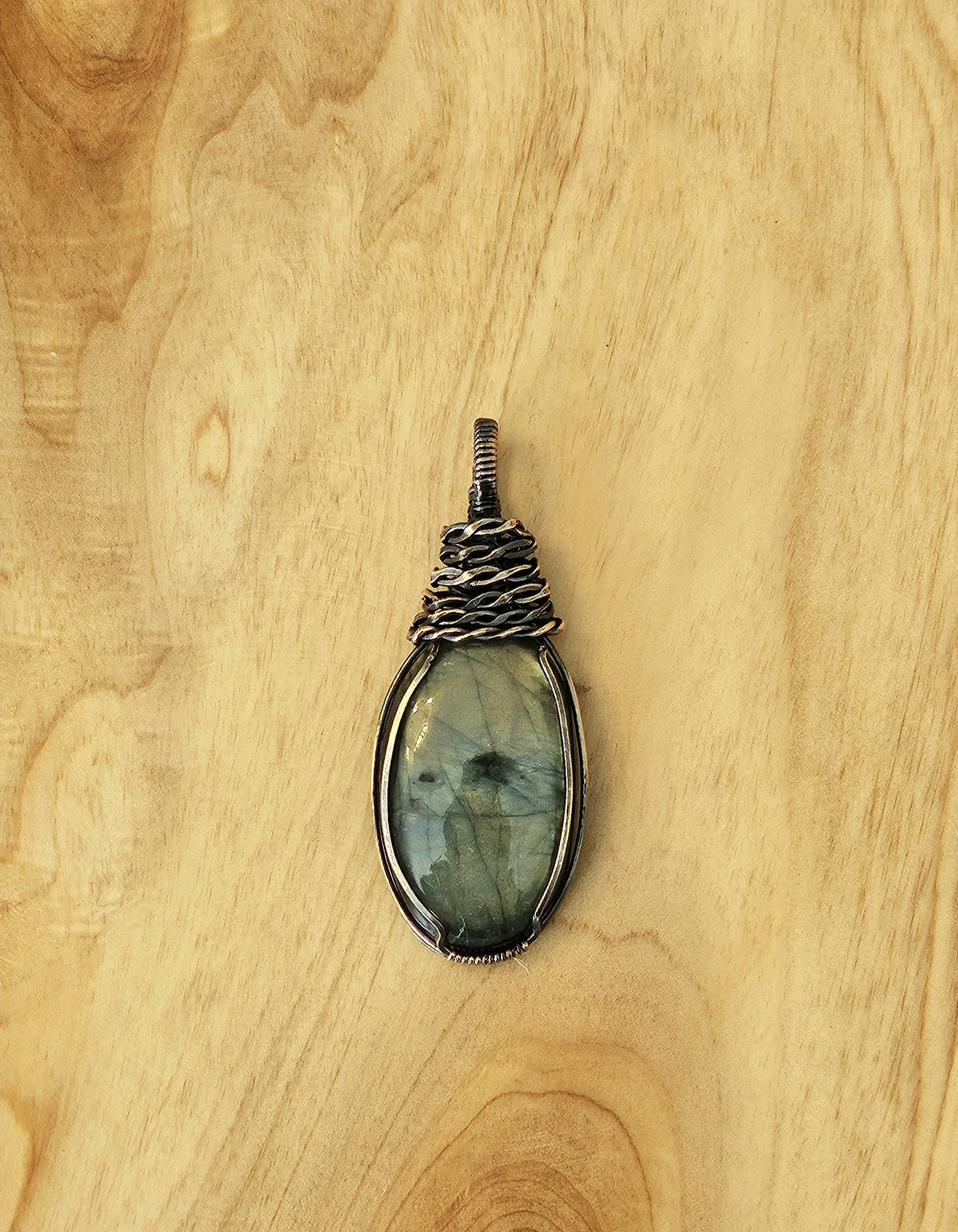 A handcrafted wire-wrapped pendant with an oval-shaped greenish, semi-transparent stone, resting on a light wooden surface.