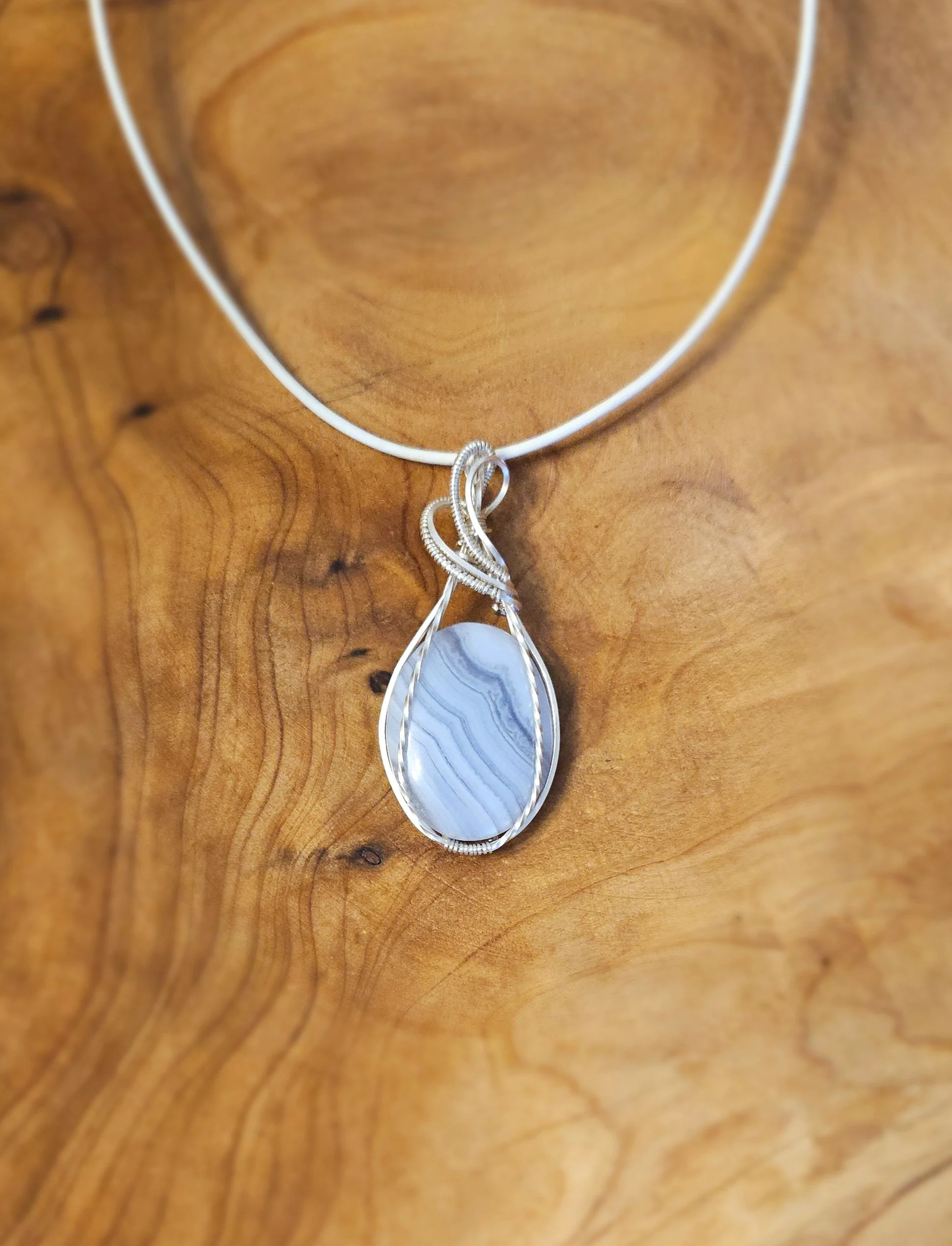 Silver necklace with a large teardrop-shaped blue and gray striped stone pendant hanging from a fancy silver chain, displayed against a wooden background.
