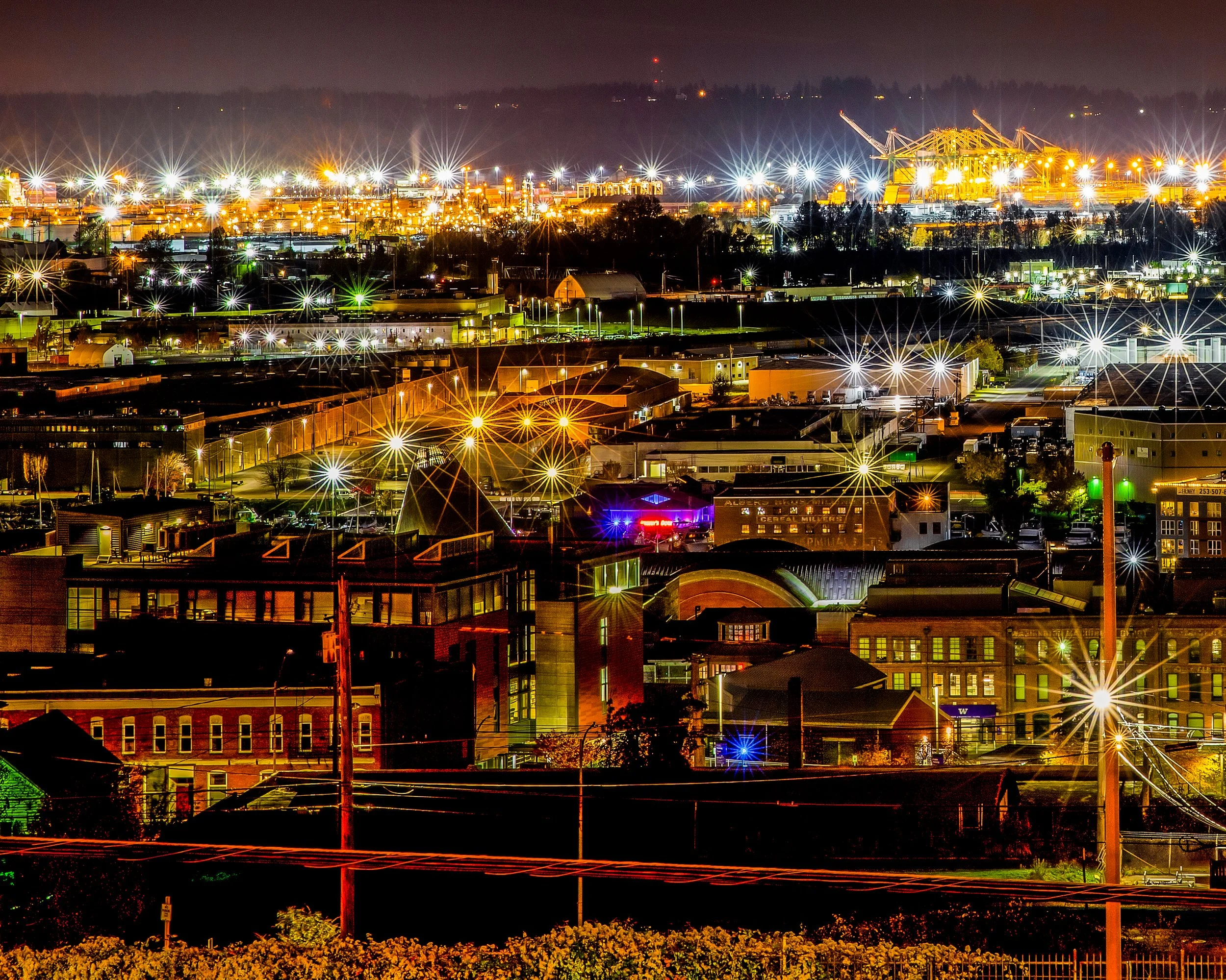 Nighttime cityscape with illuminated buildings, industrial structures, and bright streetlights, creating starburst effects across the scene.